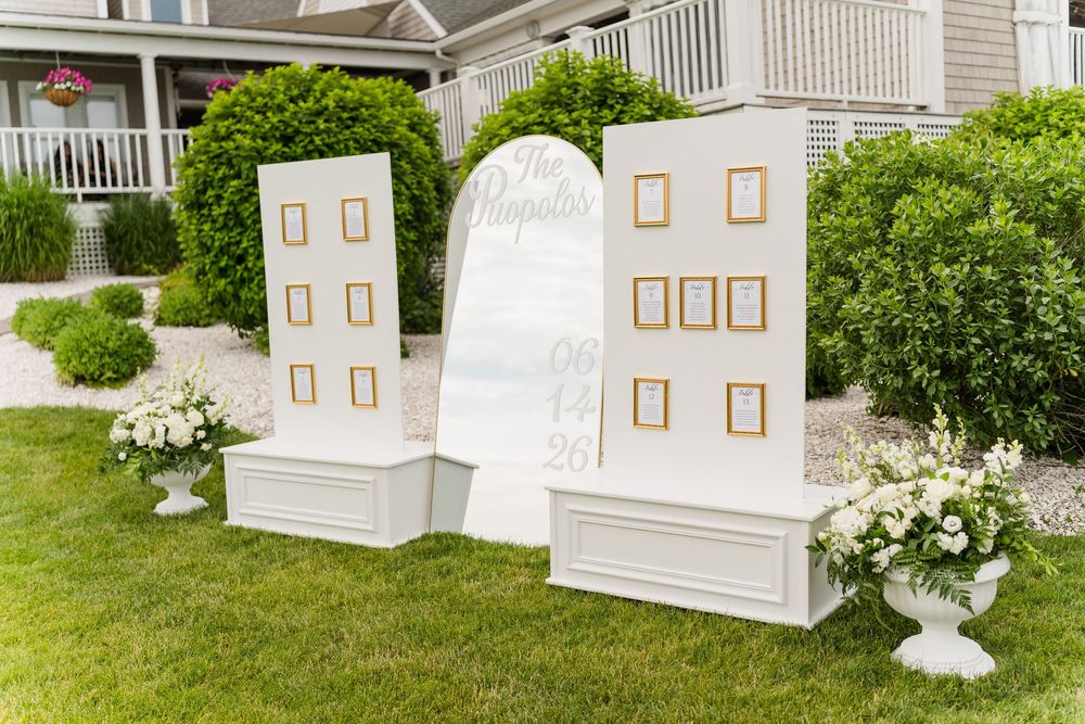 Elegant white wedding seating chart display with gold frames on lush green lawn, adorned with white floral arrangements.