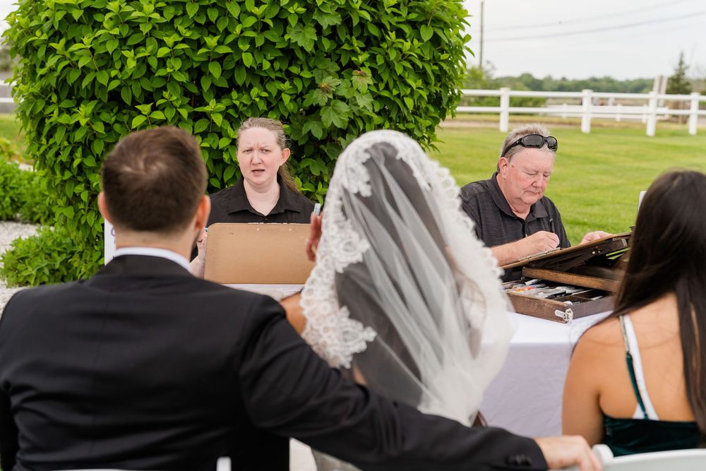 Bride and groom sit across from guests at an outdoor wedding reception table on a sunny day.