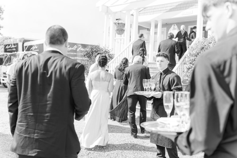 Black and white photo of a wedding party outside a venue, with a waiter serving champagne glasses to guests.