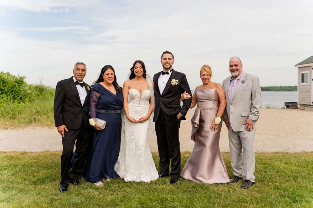 Wedding party of six posing outdoors near waterfront, bride in white gown, groom in tuxedo, guests in formal attire.