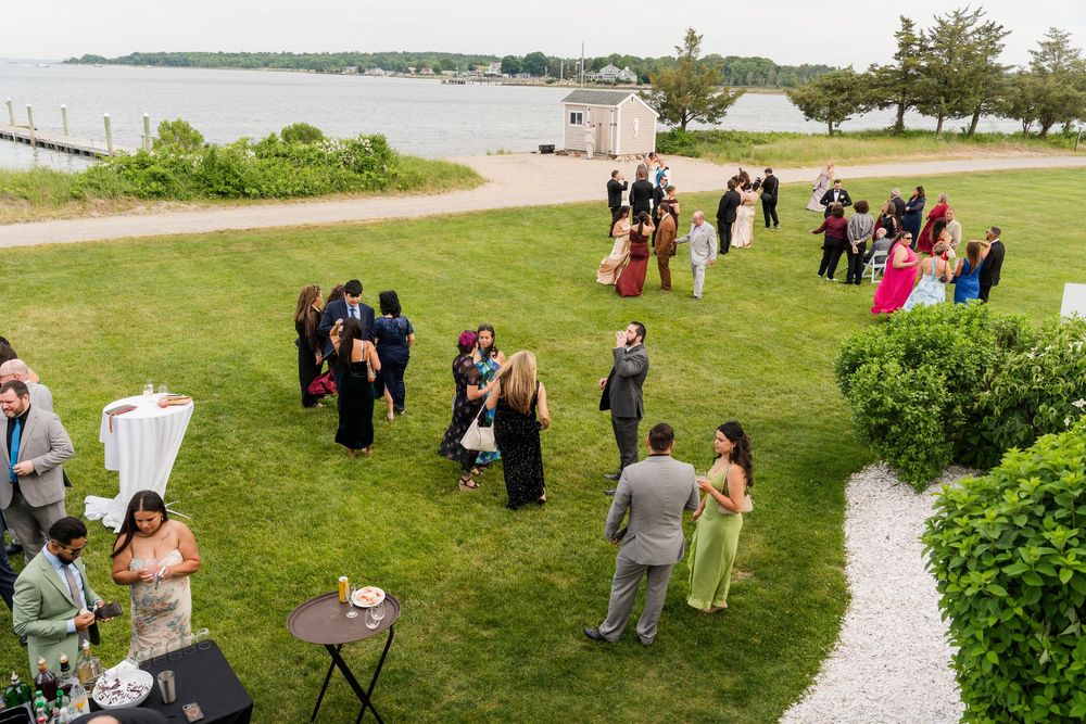 Aerial view of wedding guests mingling on a waterfront lawn during an outdoor cocktail hour reception.