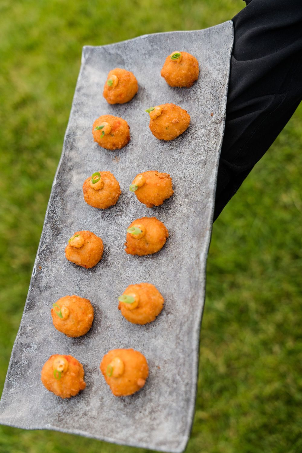 Waiter holding a metal tray with golden fried croquettes garnished with green onion, served on a grass background.