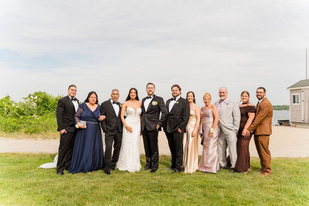 Wedding party posing outdoors on green lawn near waterfront, bride in white gown with groomsmen in tuxedos and bridesmaids in navy and gold.