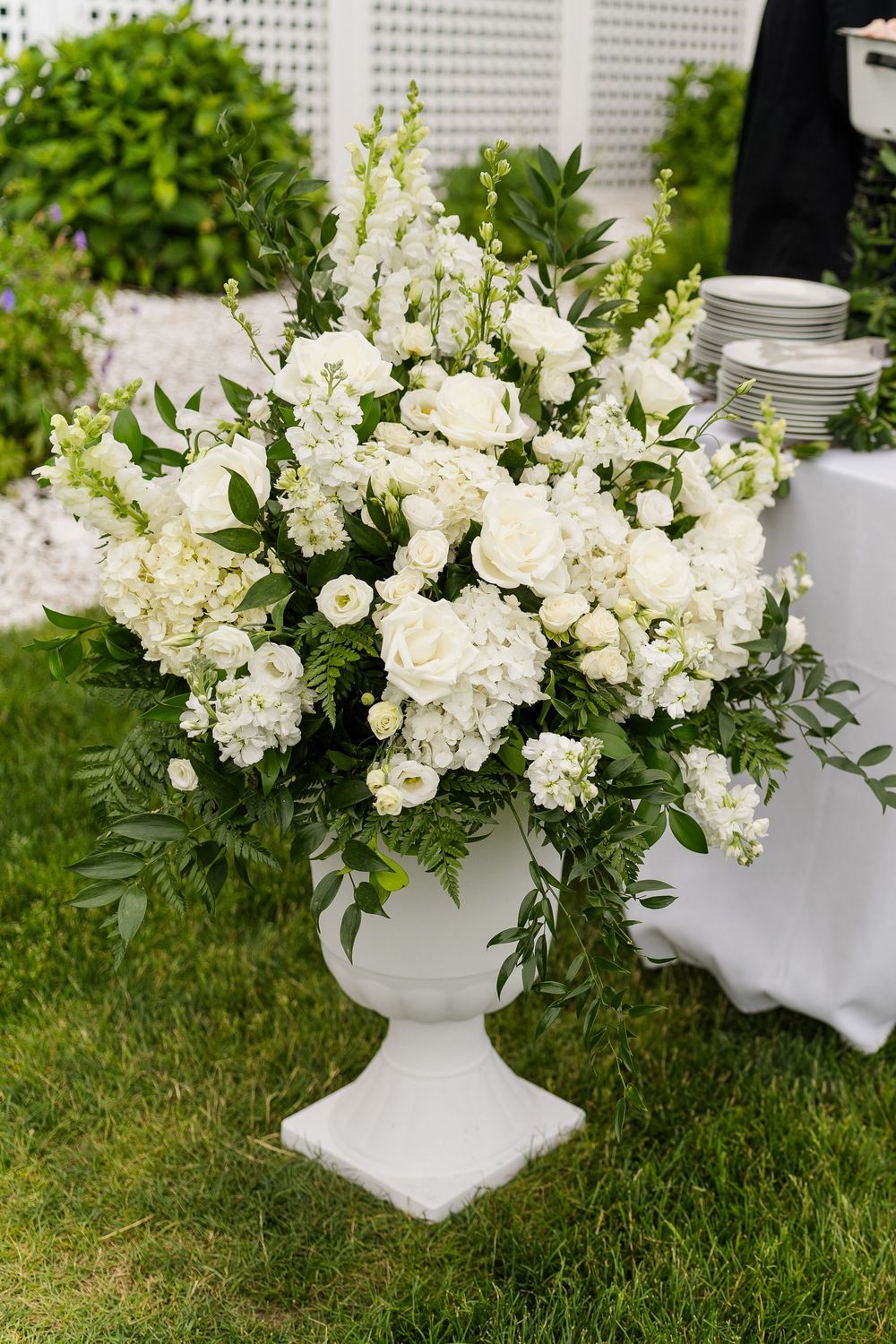 Elegant white floral arrangement in a classic white urn vase on green grass at an outdoor garden wedding event.