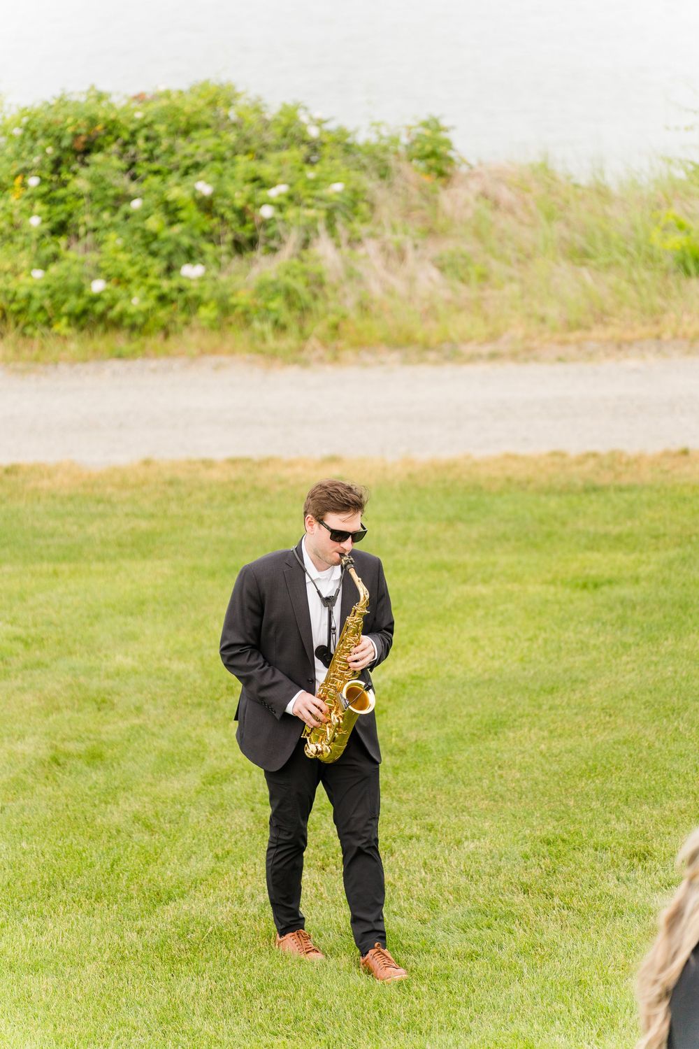 Man in black suit playing saxophone outdoors on green grass at a wedding ceremony.
