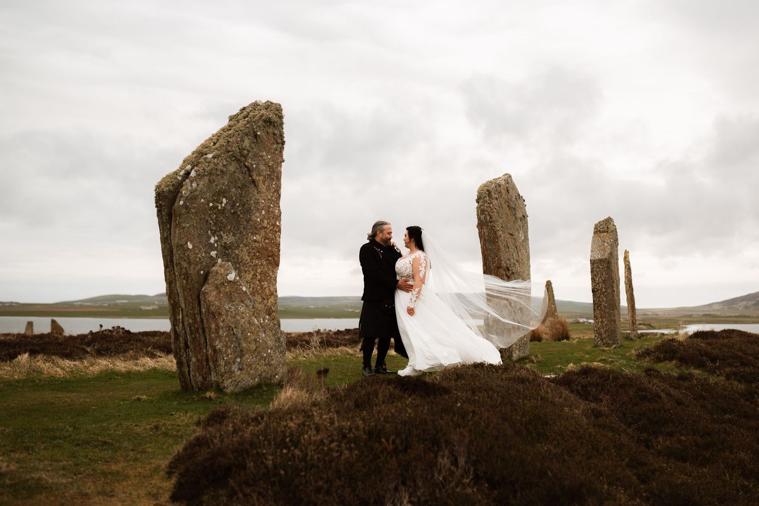 Wedding couple poses among ancient standing stones on a cloudy day in Scotland's rugged landscape.