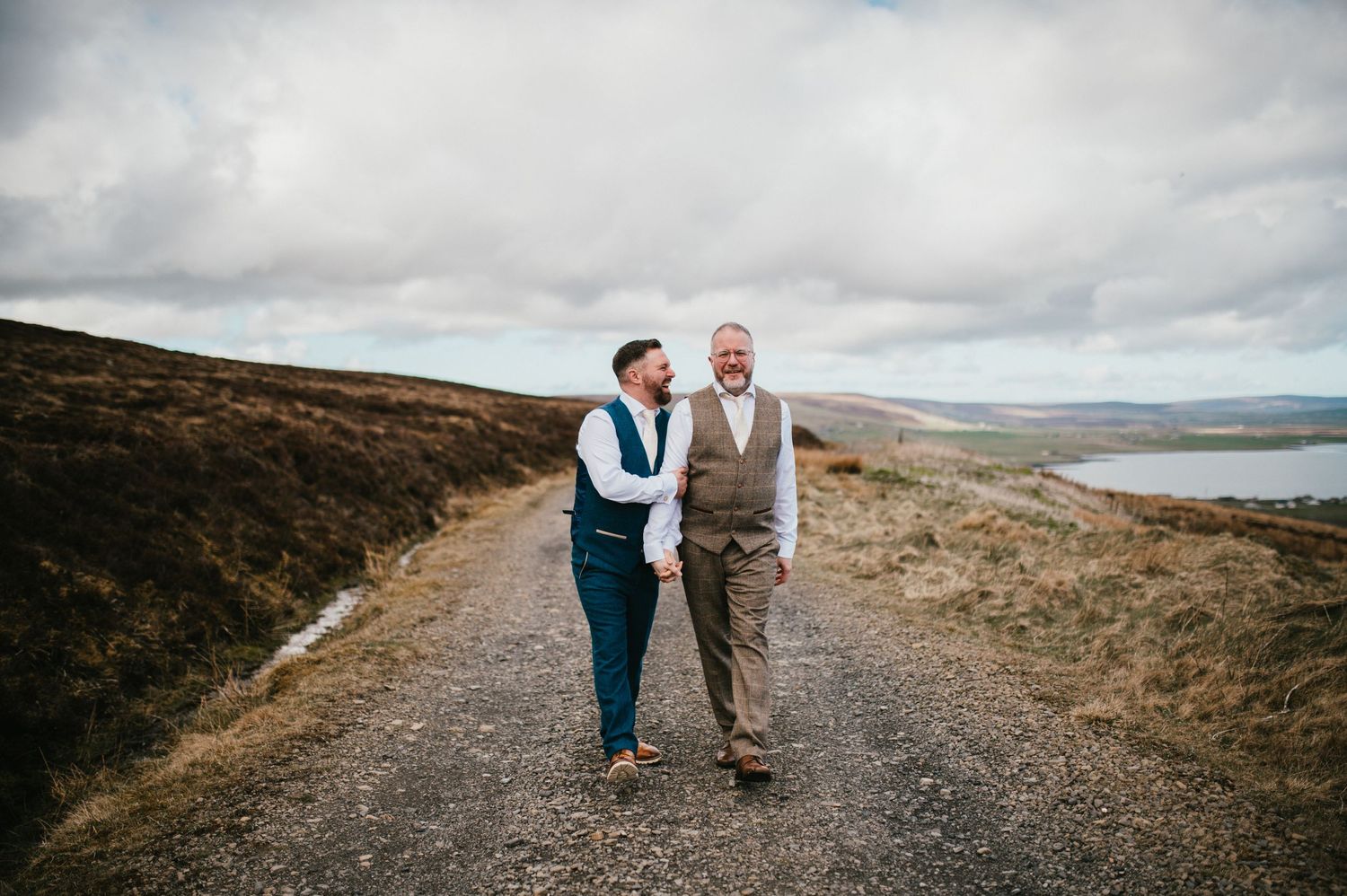 Two people walk hand in hand along a scenic gravel path overlooking a distant lake in the Scottish highlands.