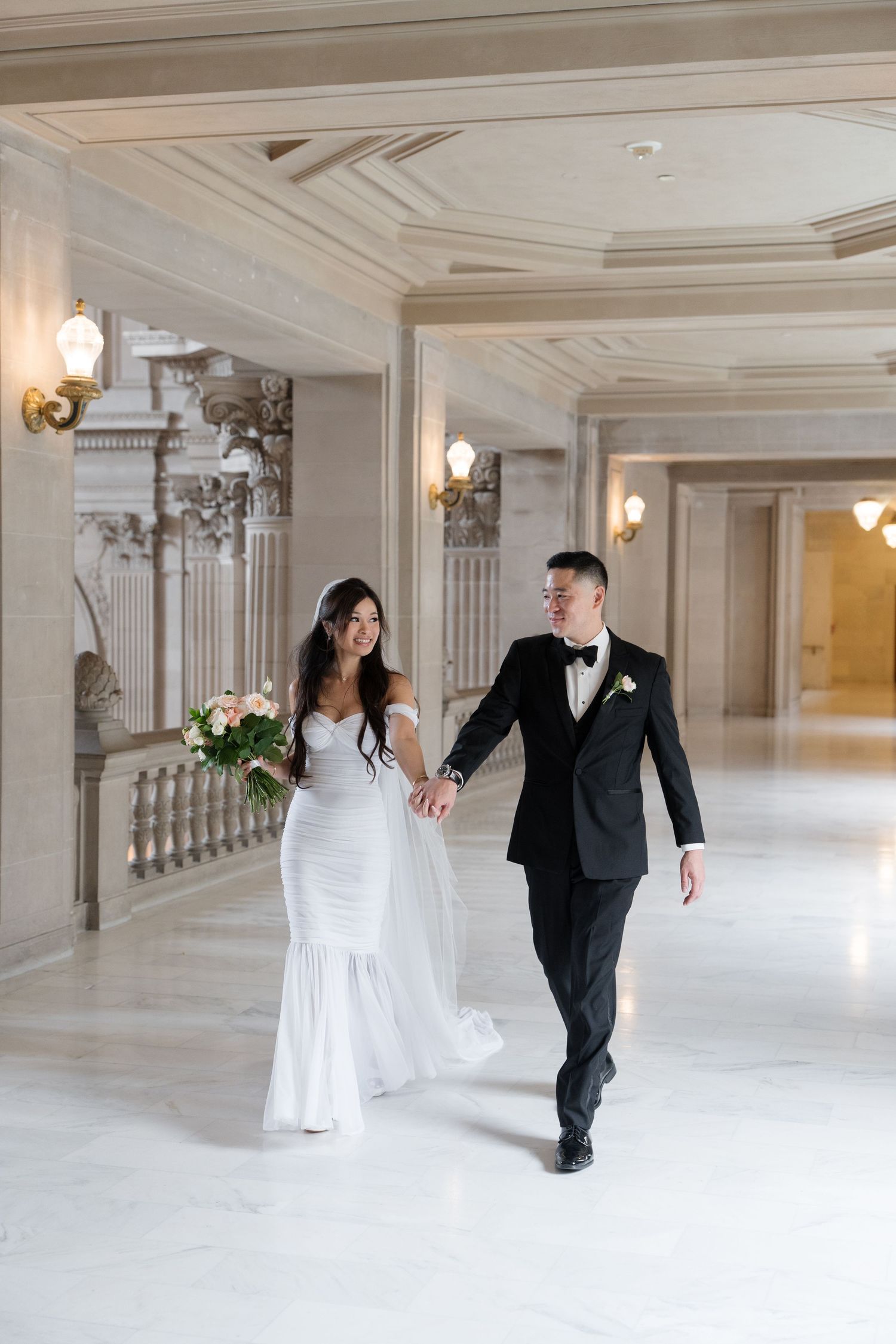A bride and groom walk hand in hand through an elegant marble hall, she in a white gown holding flowers, he in a black tuxedo.