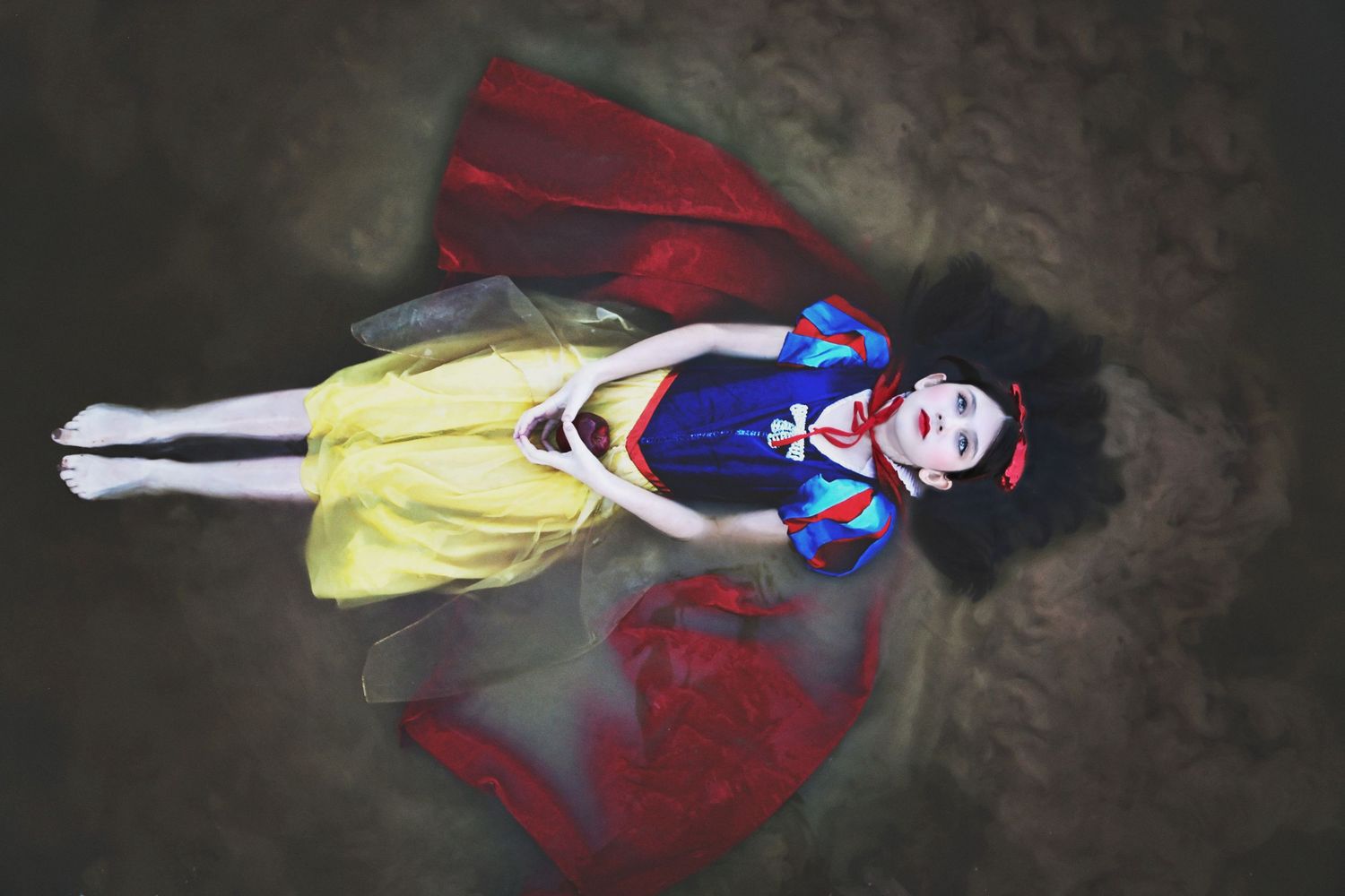 Woman in a Snow White costume with a blue bodice and yellow skirt lies on the ground with hands clasped, a red cape spread around her.