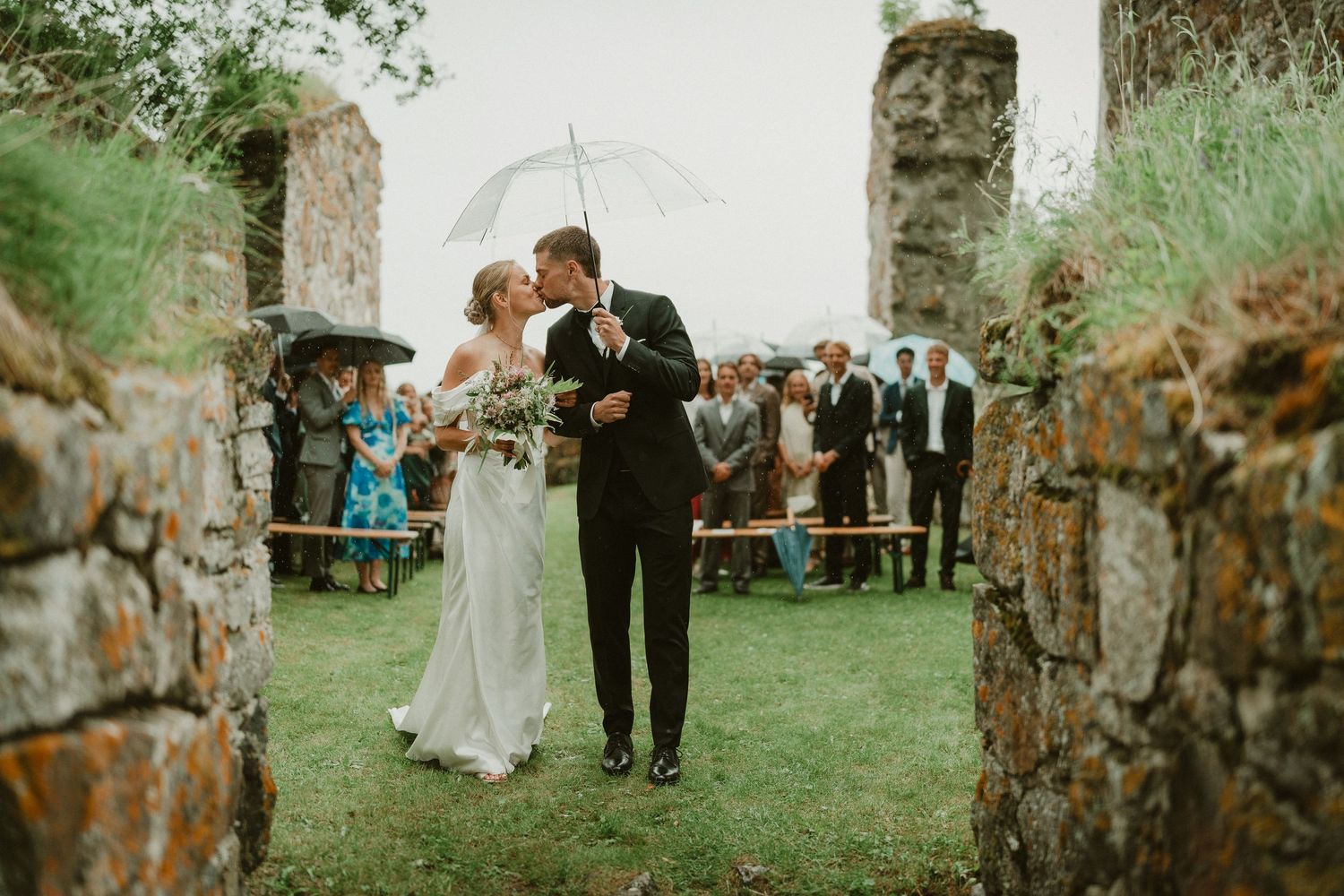 Bride and groom share a kiss under a clear umbrella at an outdoor stone ruins wedding ceremony on a rainy day.