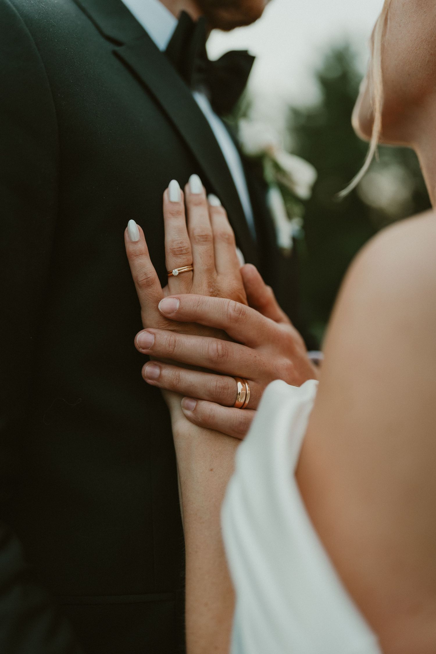Bride and groom displaying gold wedding rings on their hands, bride in white dress touching groom's black suit sleeve.
