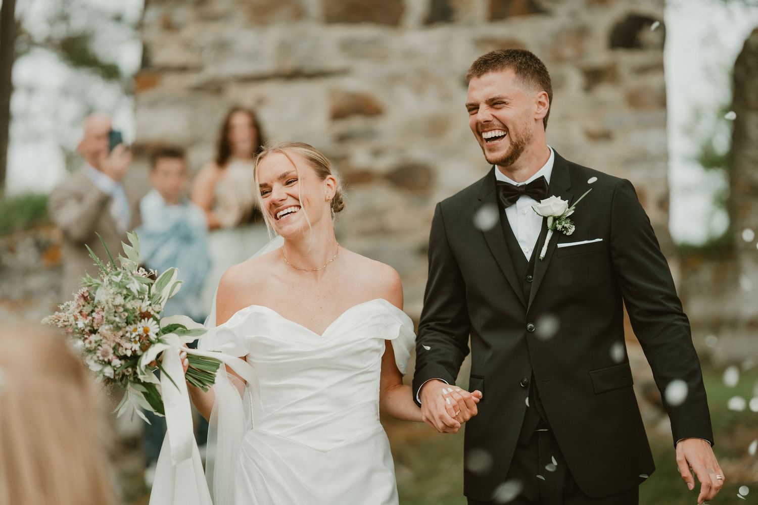 Joyful newlyweds walk hand in hand during outdoor wedding ceremony recessional with confetti and guests cheering.