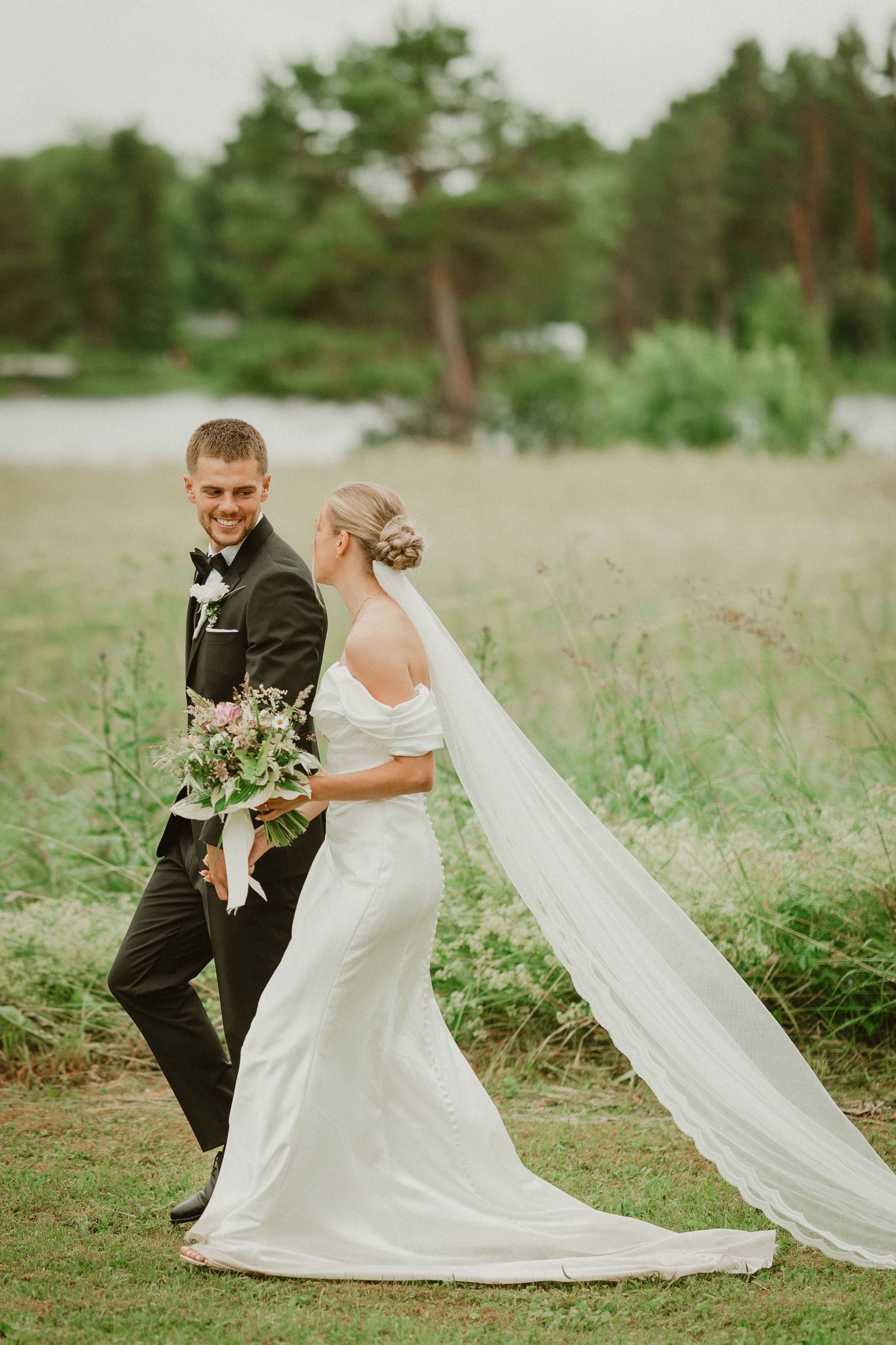 A smiling groom in a black tuxedo walks with his bride in a white gown and veil through a lush green field.