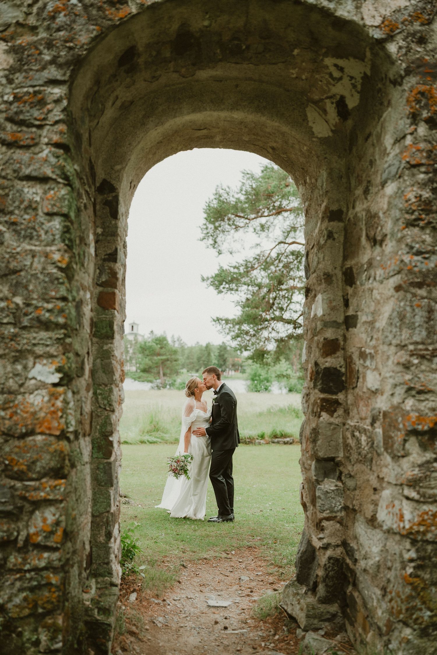 Bride and groom share a romantic moment framed by a rustic stone archway in a lush green outdoor setting.