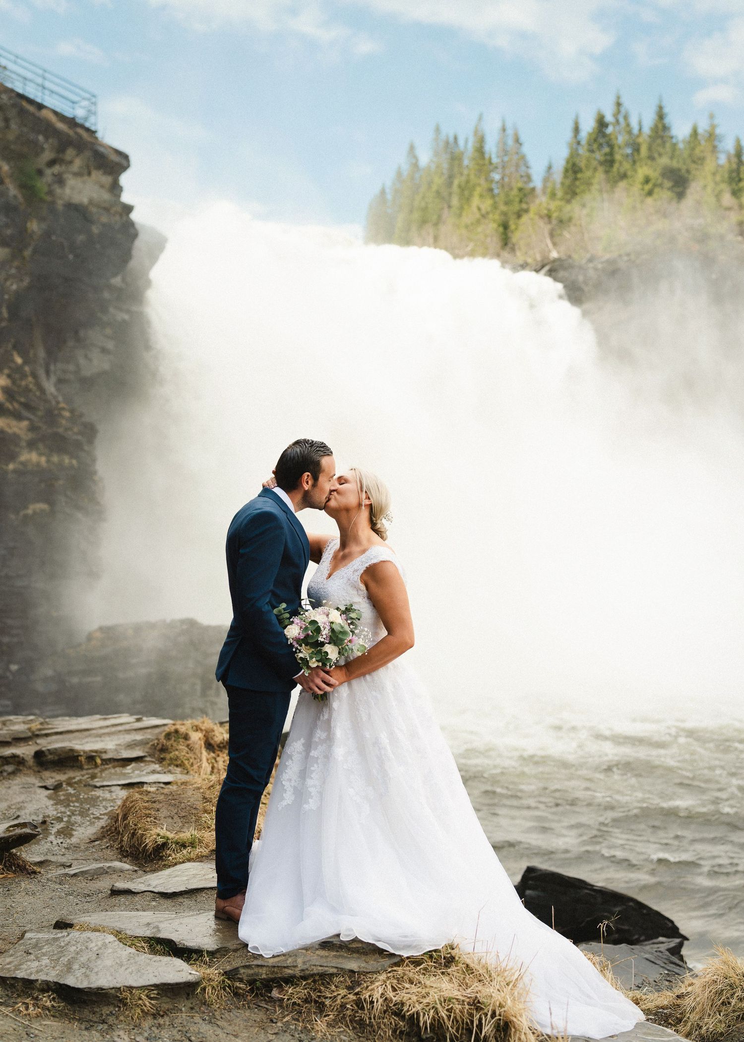 Bride and groom kissing in front of a majestic waterfall on rocky terrain, bride holding a bouquet.