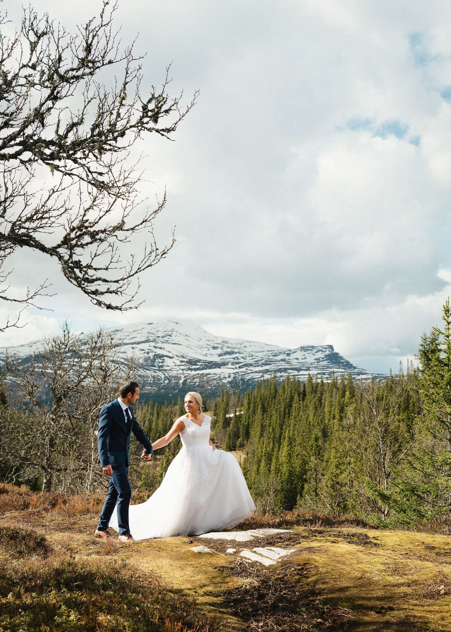 Bride and groom hold hands walking on a scenic mountain overlook with snow-capped peaks and evergreen trees.
