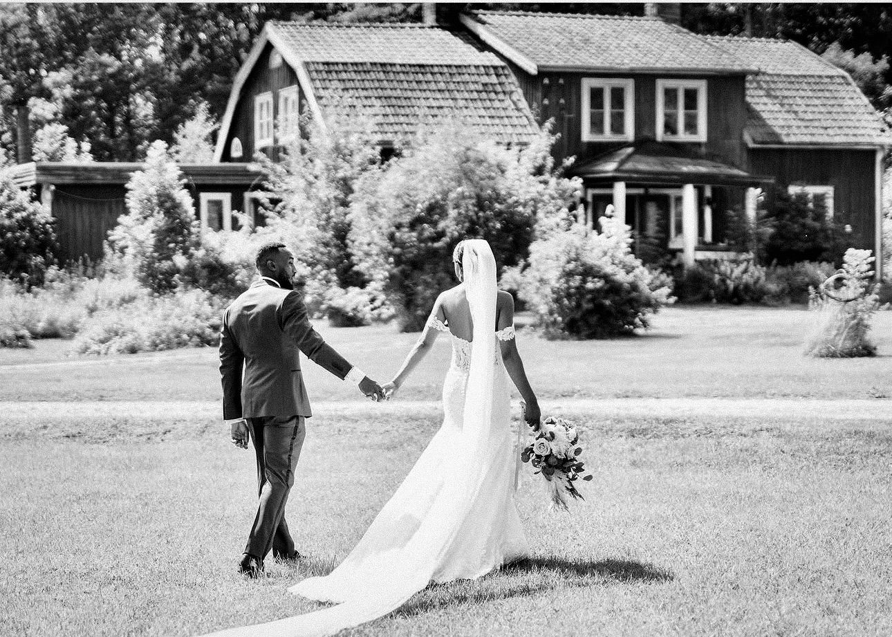 Black and white photo of a bride and groom holding hands walking toward a charming house on their wedding day.