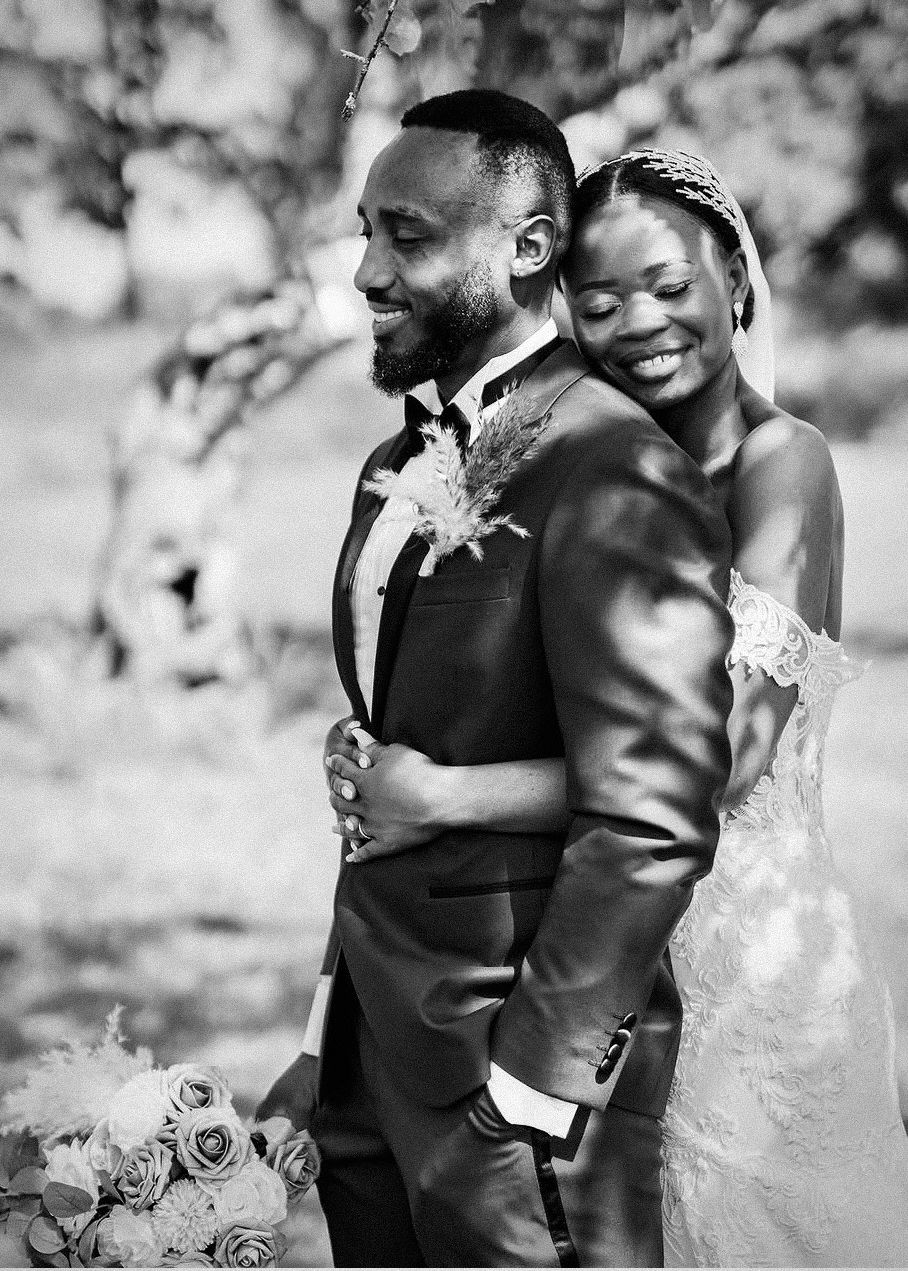 Black and white wedding photo of a joyful couple embracing outdoors, bride in lace gown holding bouquet, groom in elegant suit.