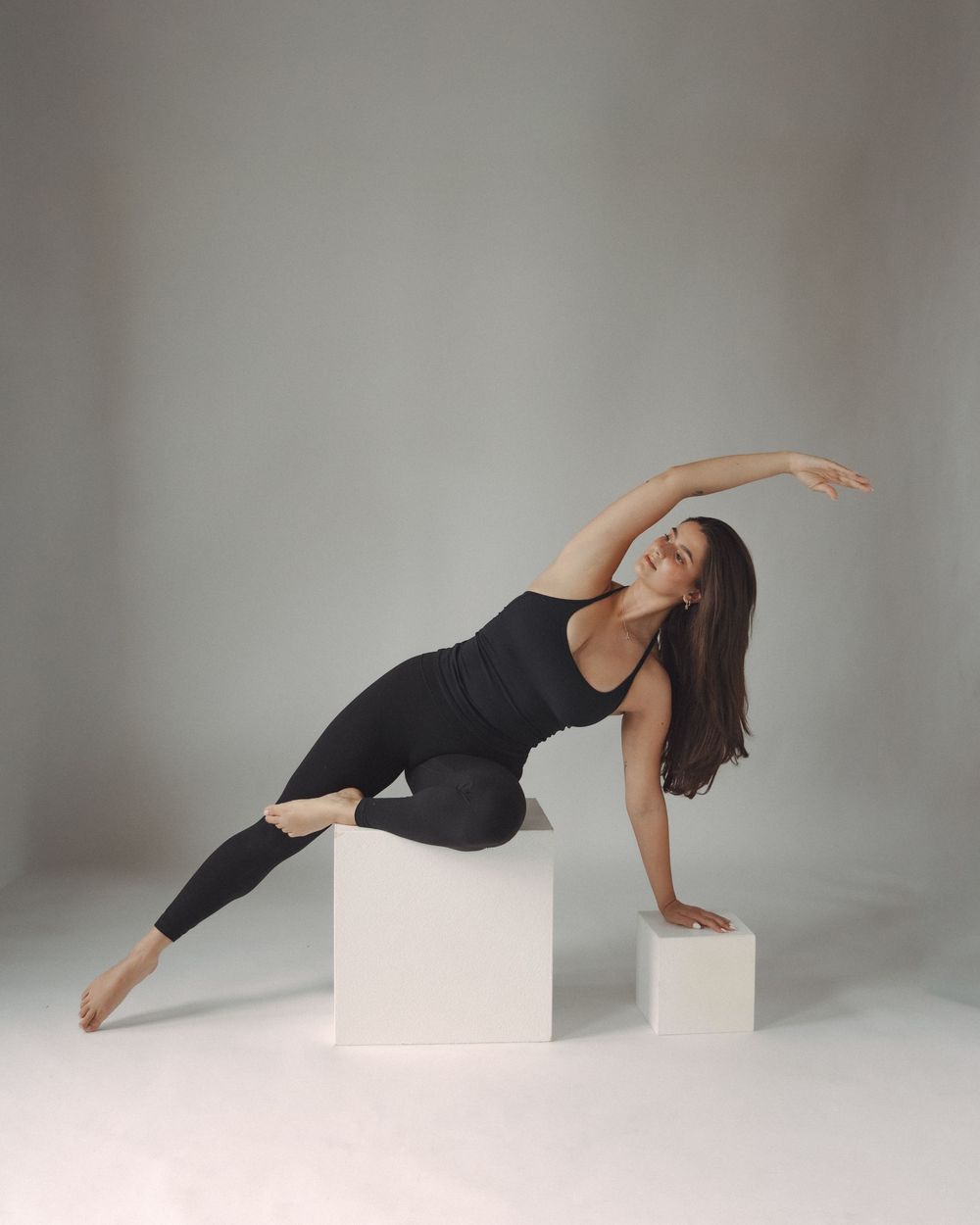 Woman in black activewear performing a side stretch pose on white cubes against a gray studio background.