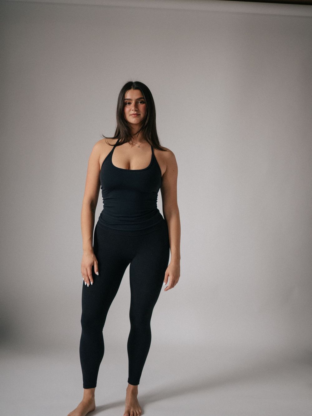 Woman in black sleeveless jumpsuit and leggings standing barefoot against a light gray background.