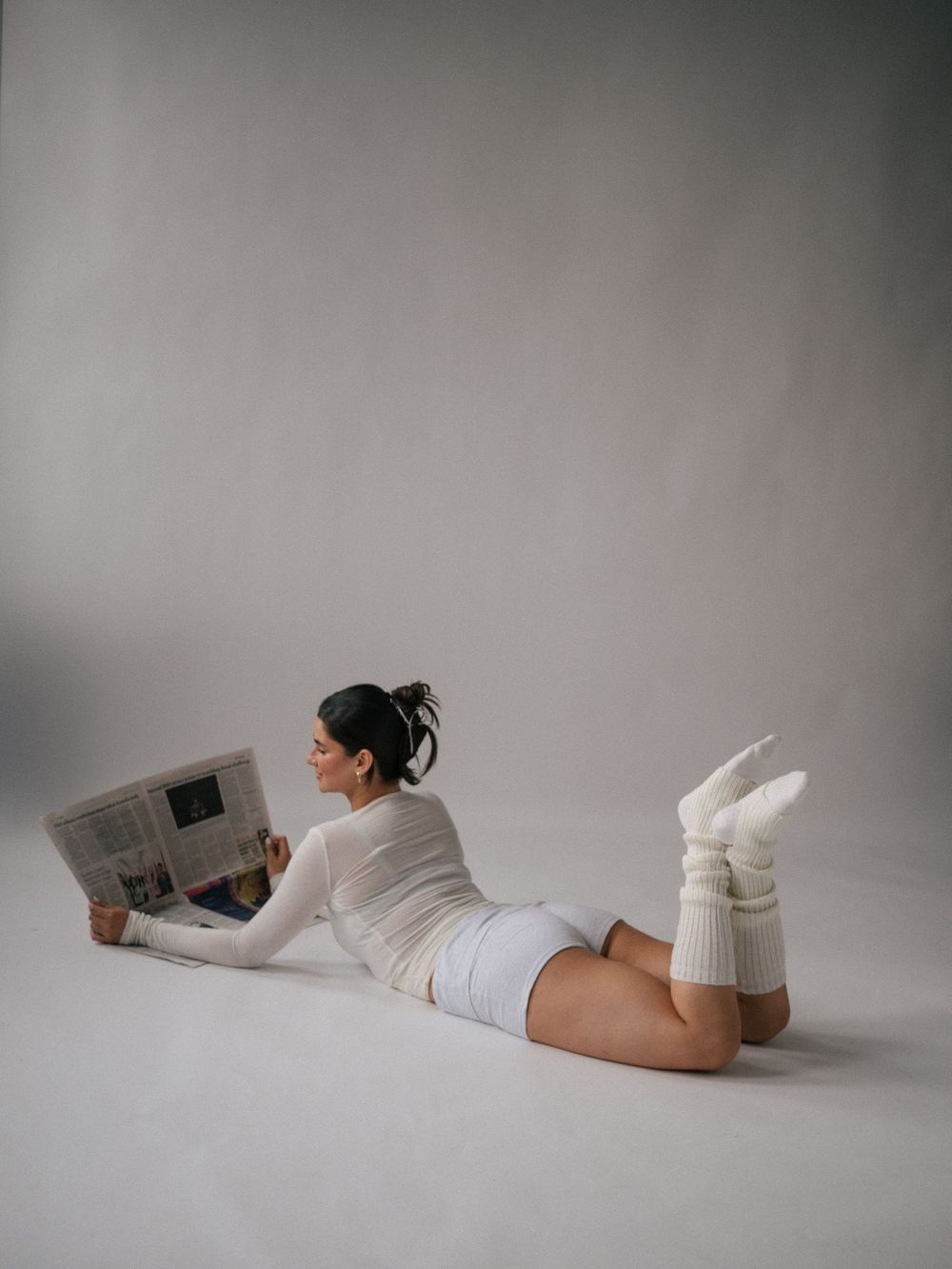 Woman in white outfit lying on floor reading newspaper, wearing chunky knit socks, studio photo.