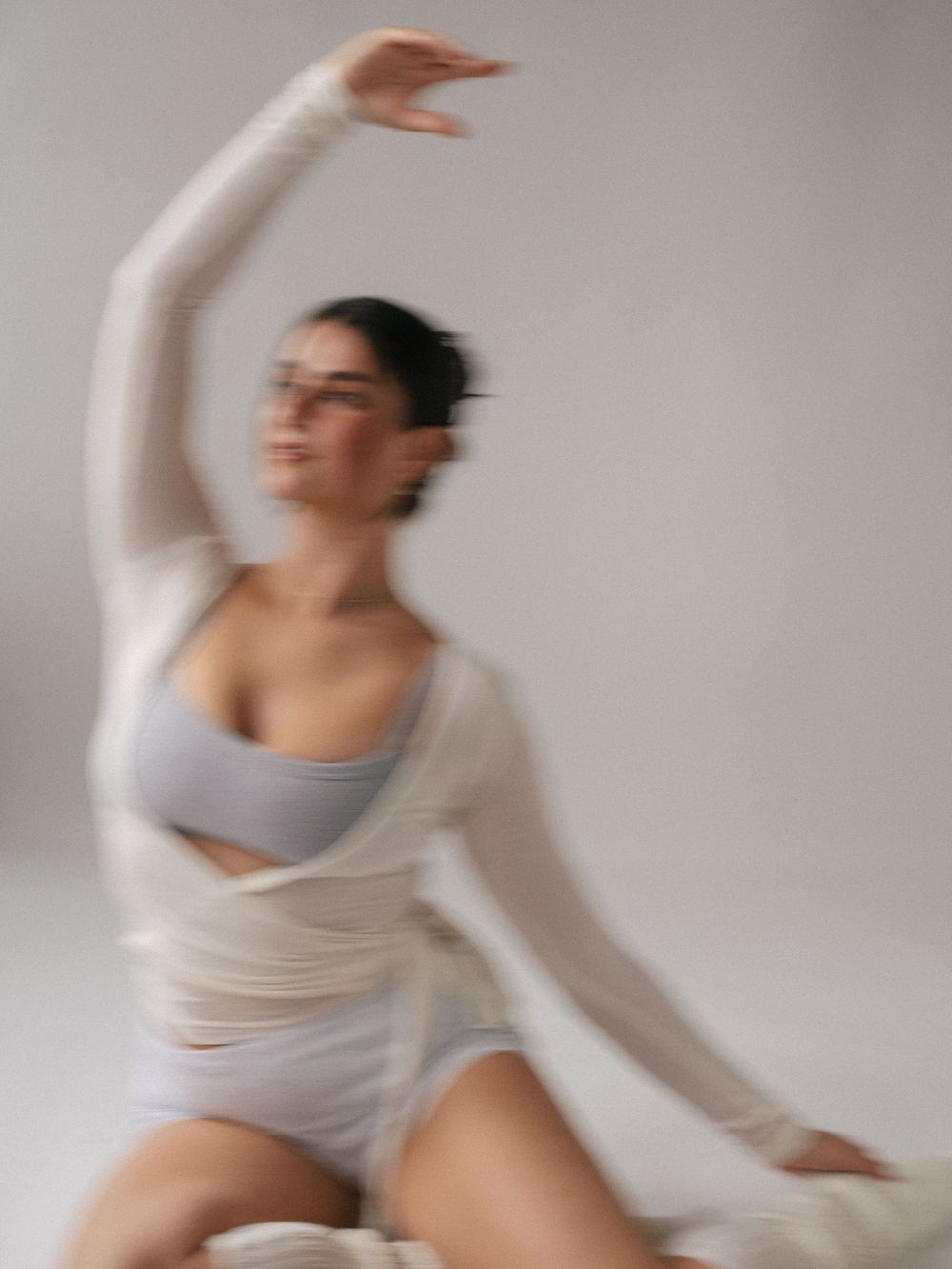 Woman in white sheer top and gray sports bra poses gracefully with arm raised in a soft-focus artistic photo.