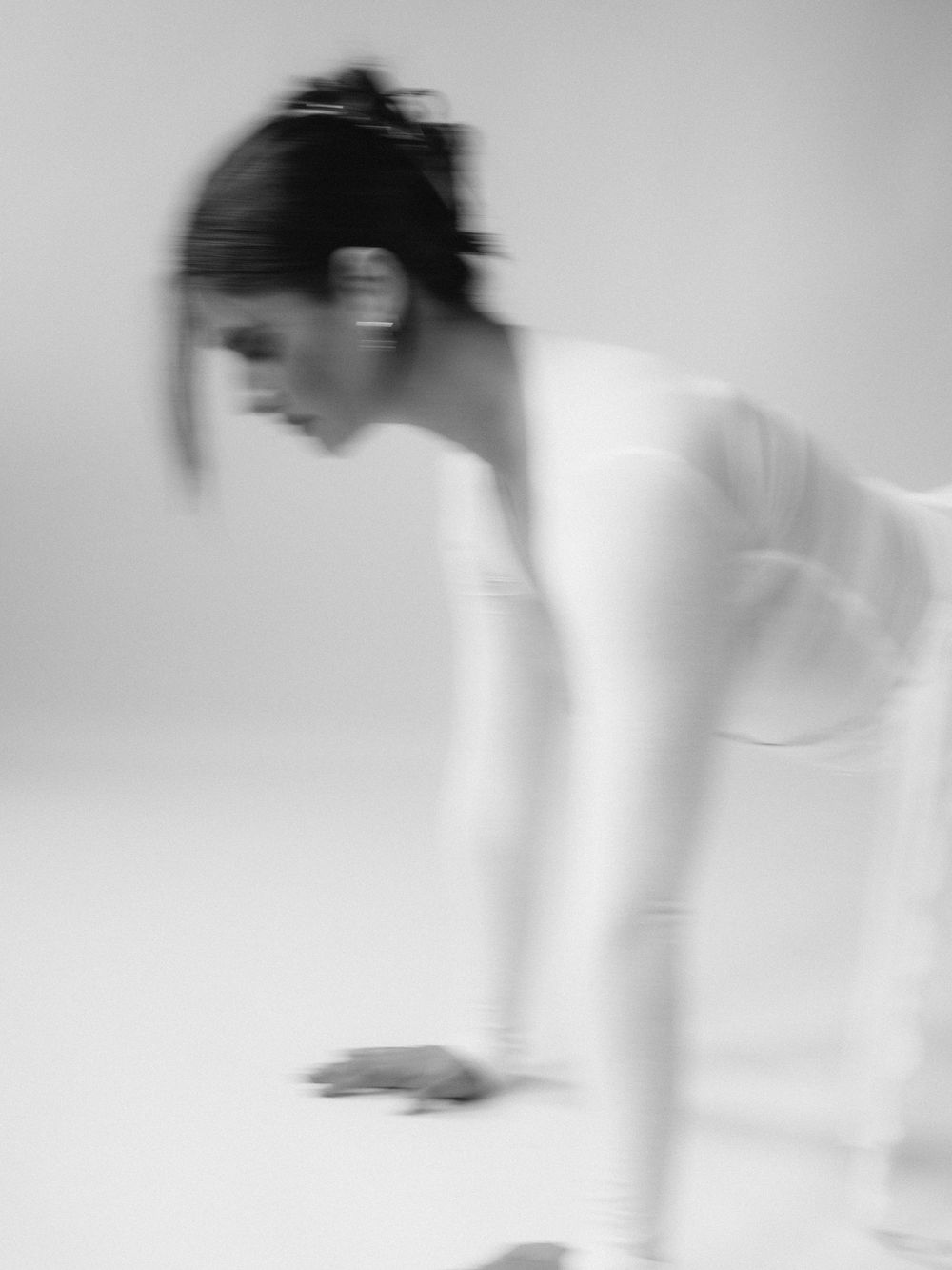 Blurred black and white photo of a woman in white bodysuit leaning forward in a dynamic dance pose.