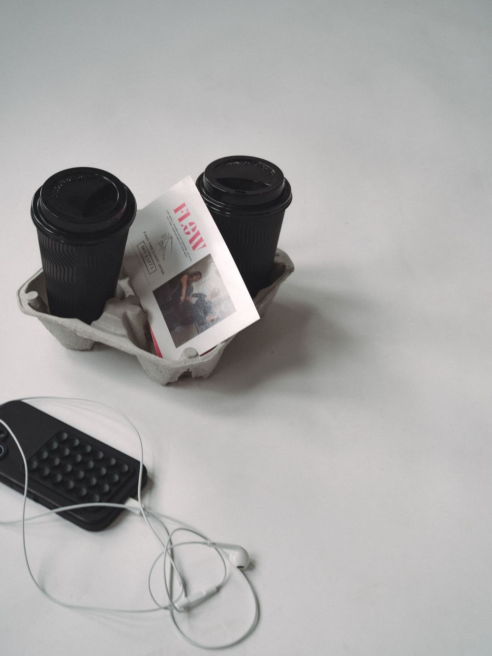 Two black coffee cups in a cardboard carrier with a brochure, beside a remote control with earphones on white background.