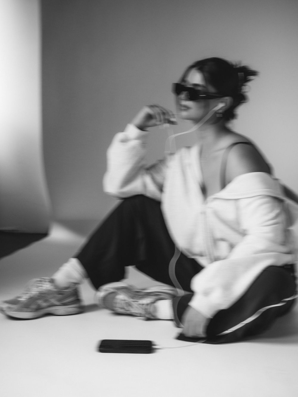 Black and white photo of a stylish woman sitting on floor, wearing sunglasses, oversized jacket, and chunky sneakers.