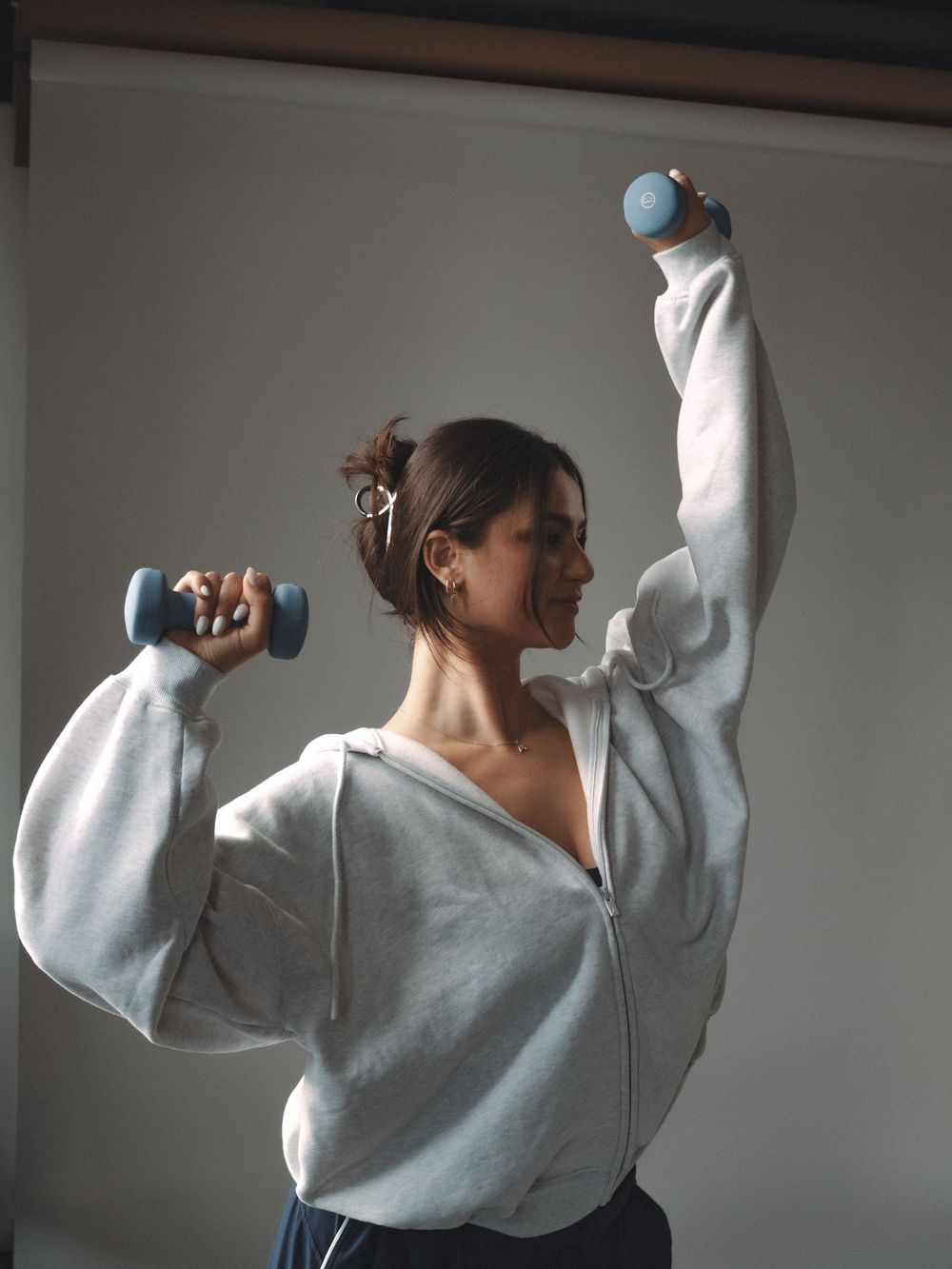 Woman in gray zip-up hoodie exercising with blue dumbbells, one arm raised overhead in a sunlit room.
