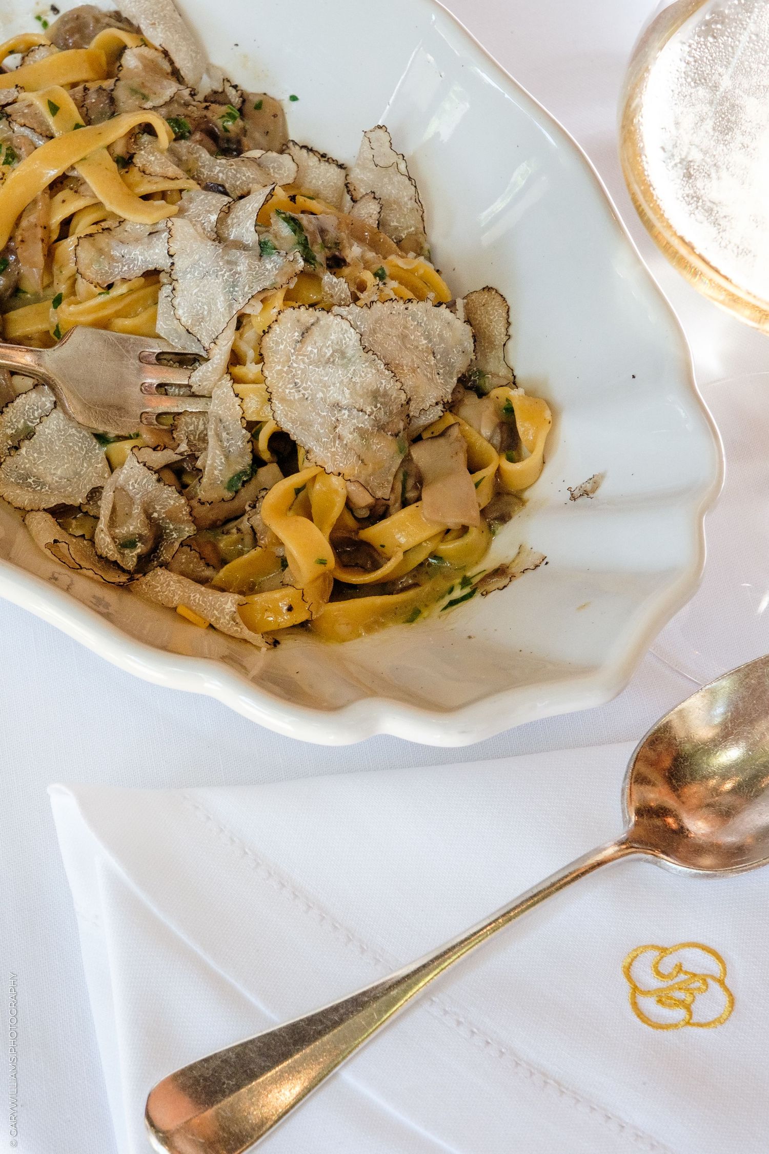 Tagliatelle pasta with shaved white truffles served in elegant white bowl with gold cutlery on white linen.