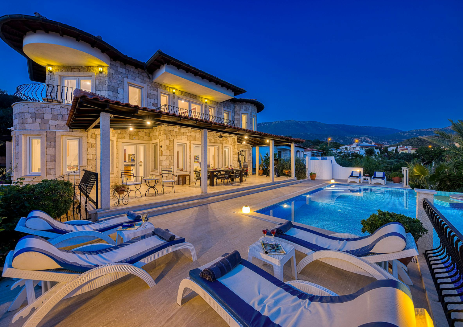 Elegant stone villa at night with lit swimming pool, lounge chairs, and warm ambient lighting on terraced patio.