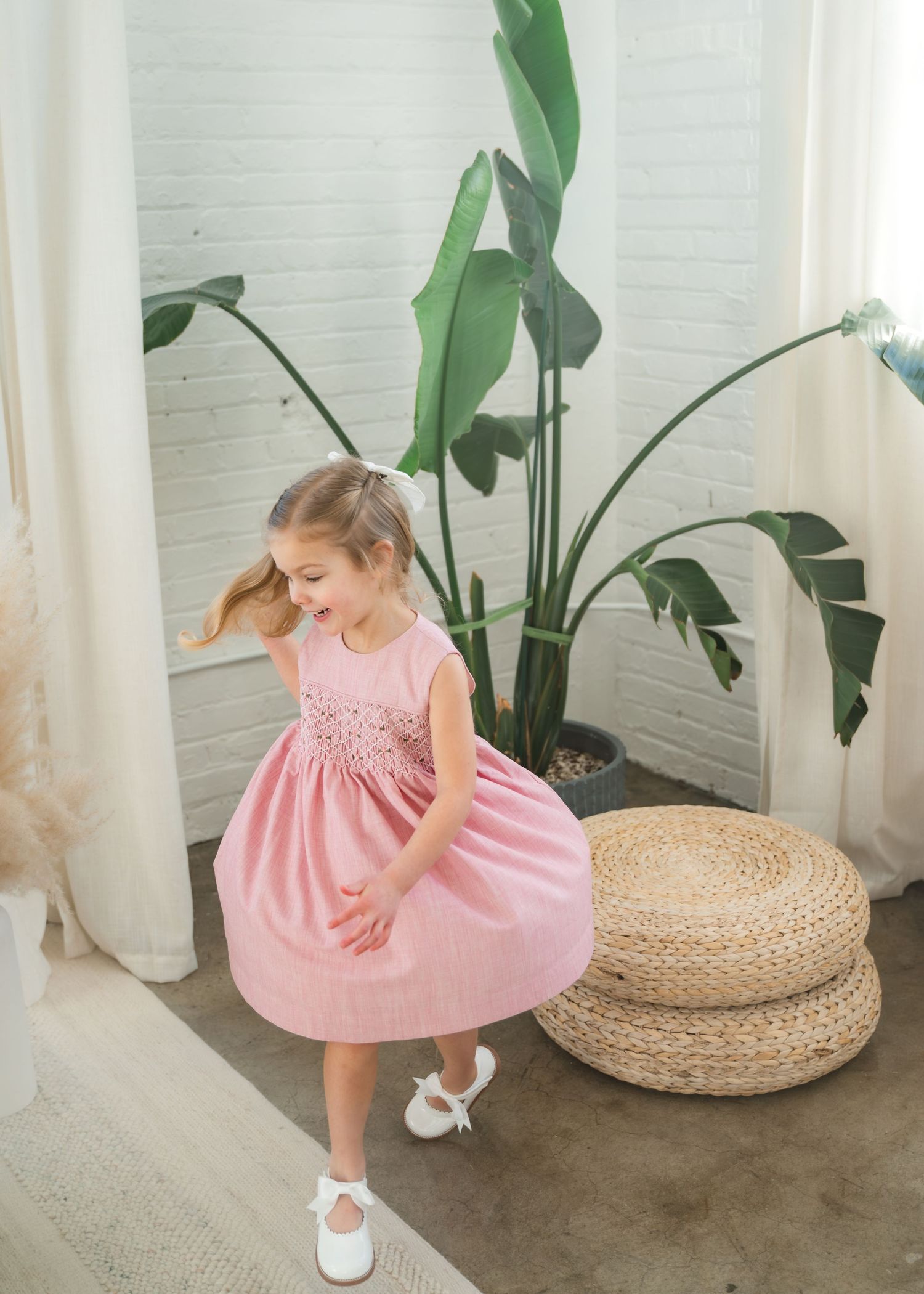 Young girl twirling in a pink smocked dress with white shoes, posing near a tropical plant indoors.