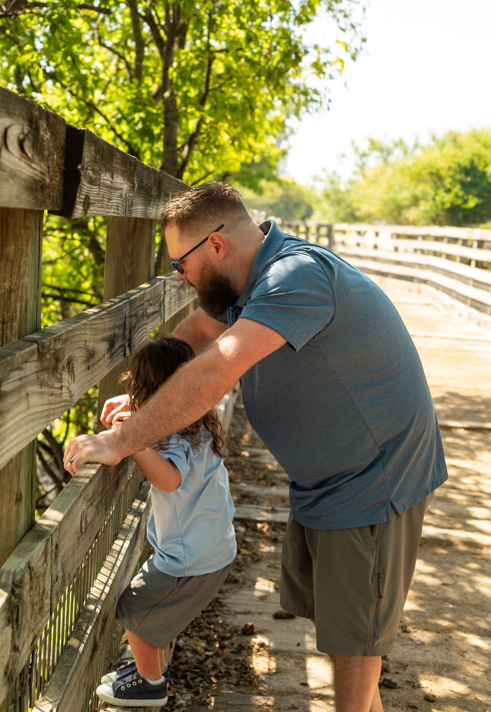 A father helps his young child look through a wooden fence on a sunny bridge surrounded by green trees.