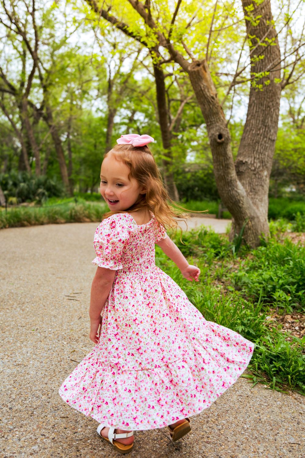 Young girl twirling in a pink floral dress with a bow in her hair on a park path surrounded by green trees.