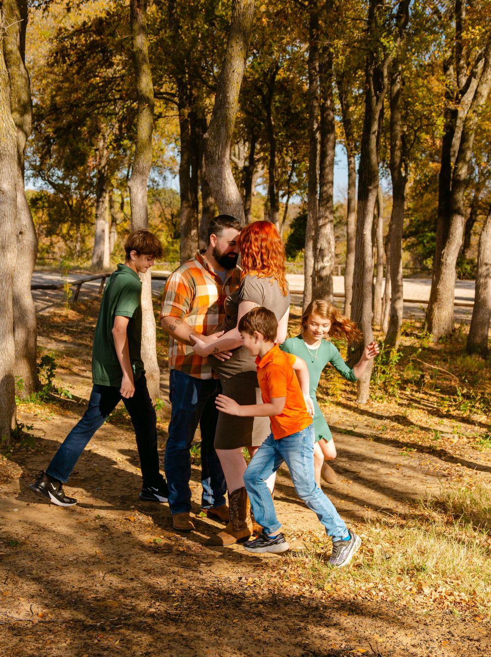 Family walking together on a sunny autumn forest path surrounded by colorful fall foliage and tall trees.