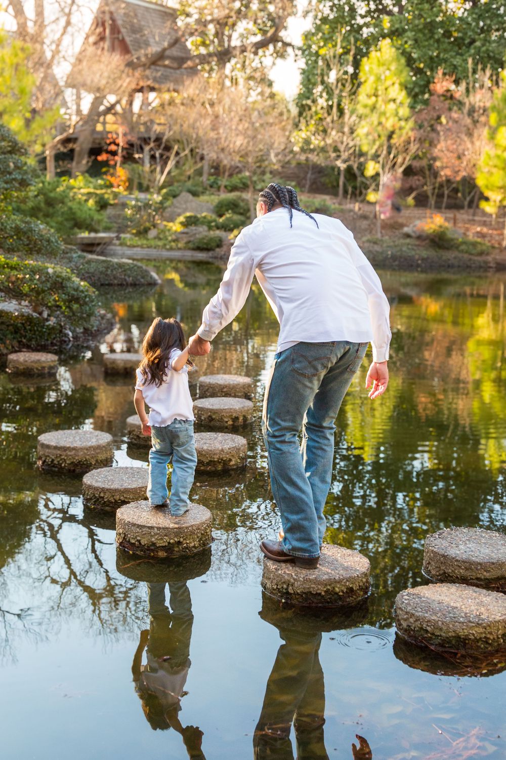Father and child holding hands while crossing stepping stones over a serene pond in a Japanese garden.