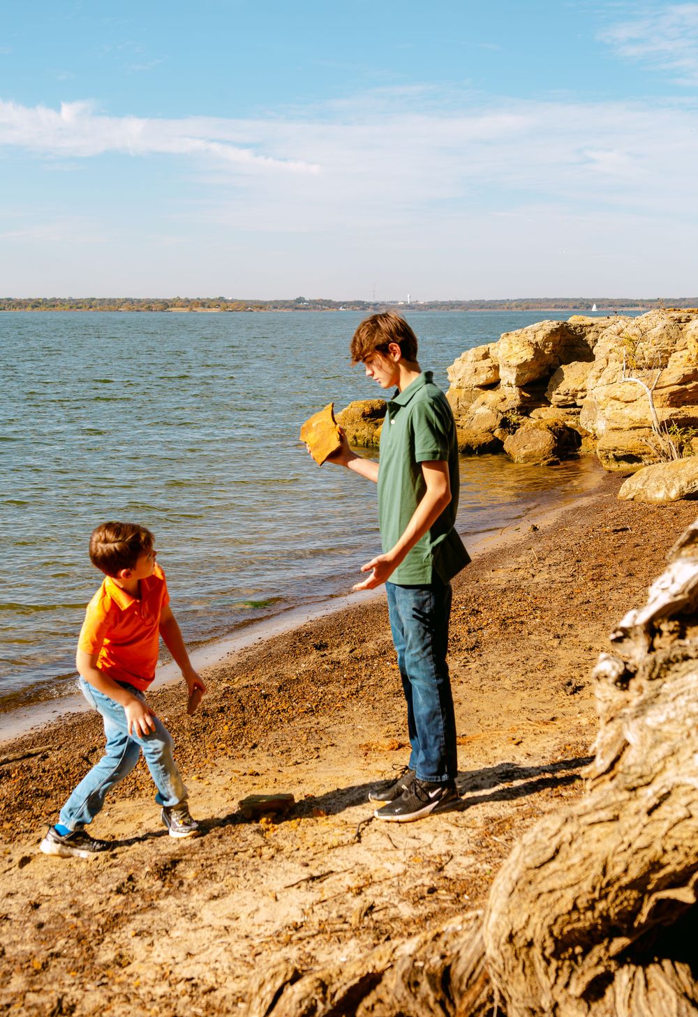 Two boys exploring a rocky shoreline, with one holding a rock near calm blue waters on a sunny day.