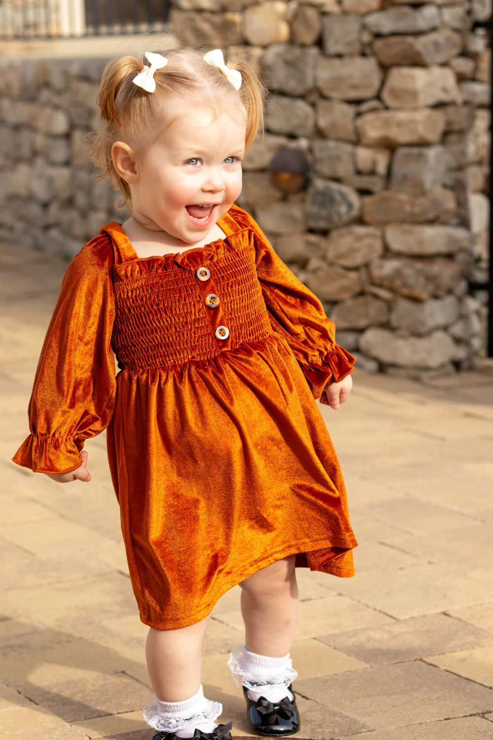 Smiling toddler girl in orange velvet dress with white hair bows and black shoes standing on brick patio.