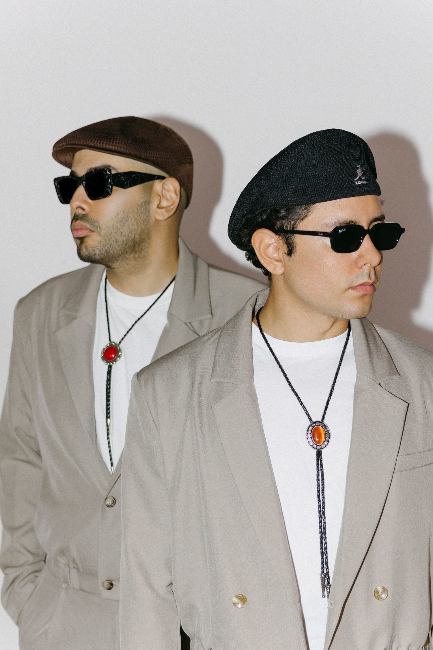 Two stylish men in beige trench coats, bolo ties, hats, and sunglasses pose against a white background.