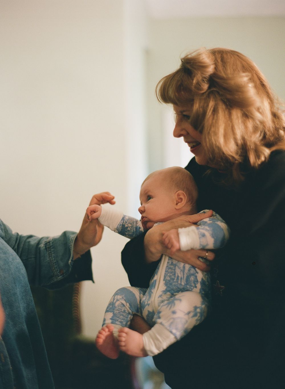 A woman with blonde hair cradles a baby in a blue onesie while someone offers the infant a bottle.