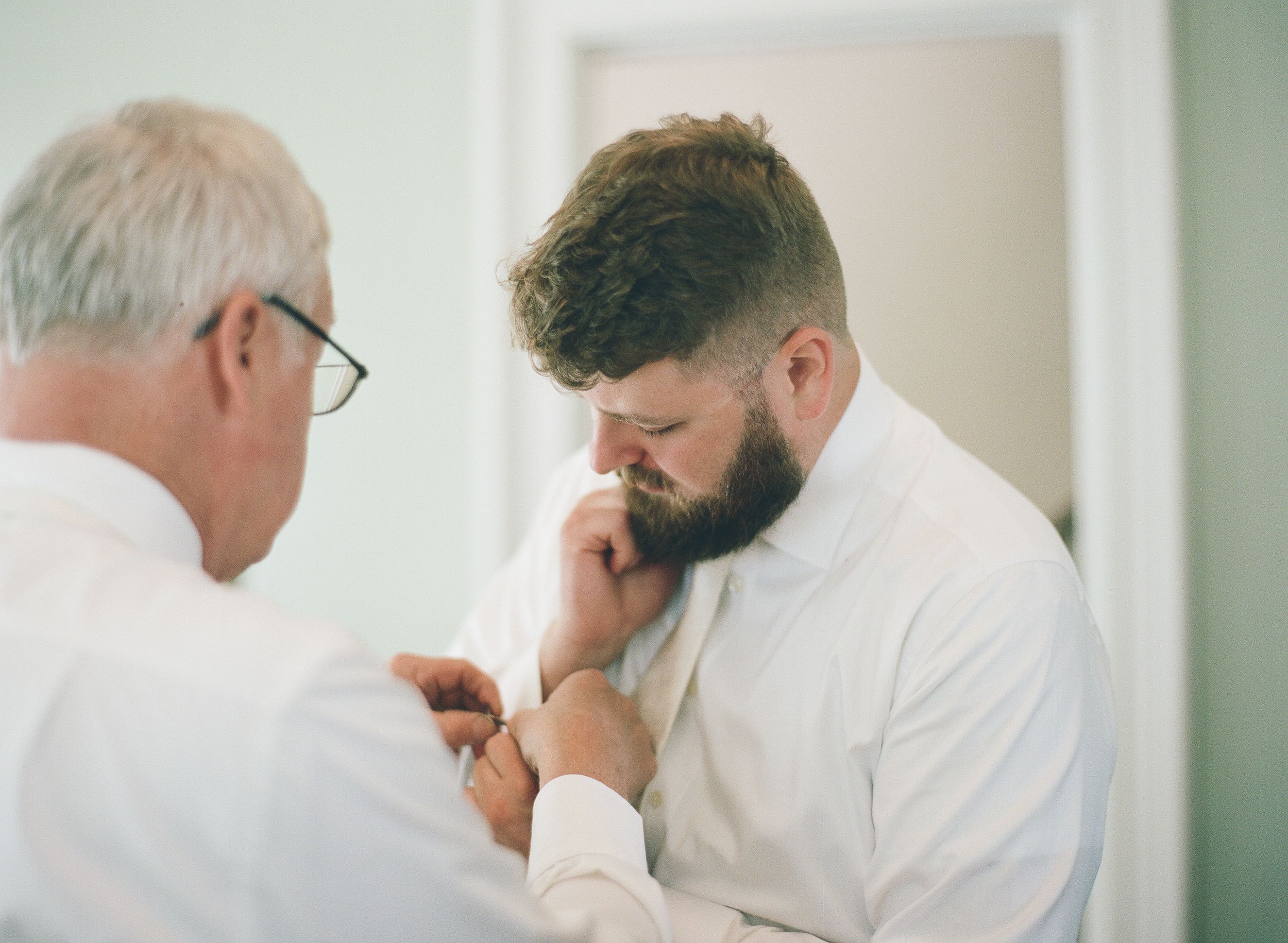 Groom in white shirt gets cufflinks fastened by older man before wedding ceremony.