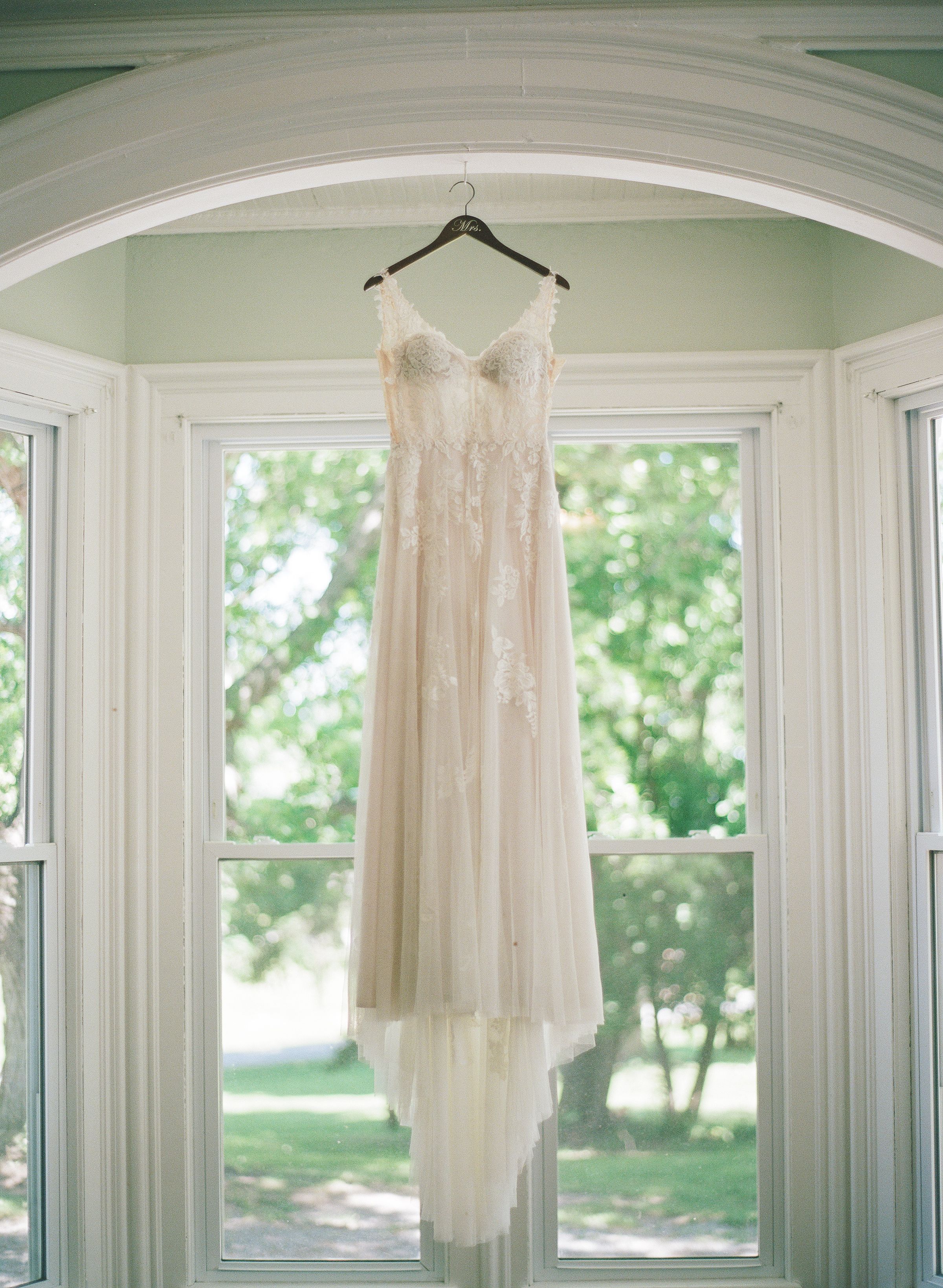Elegant lace wedding dress hanging on a hanger in a bright bay window with green trees visible outside.