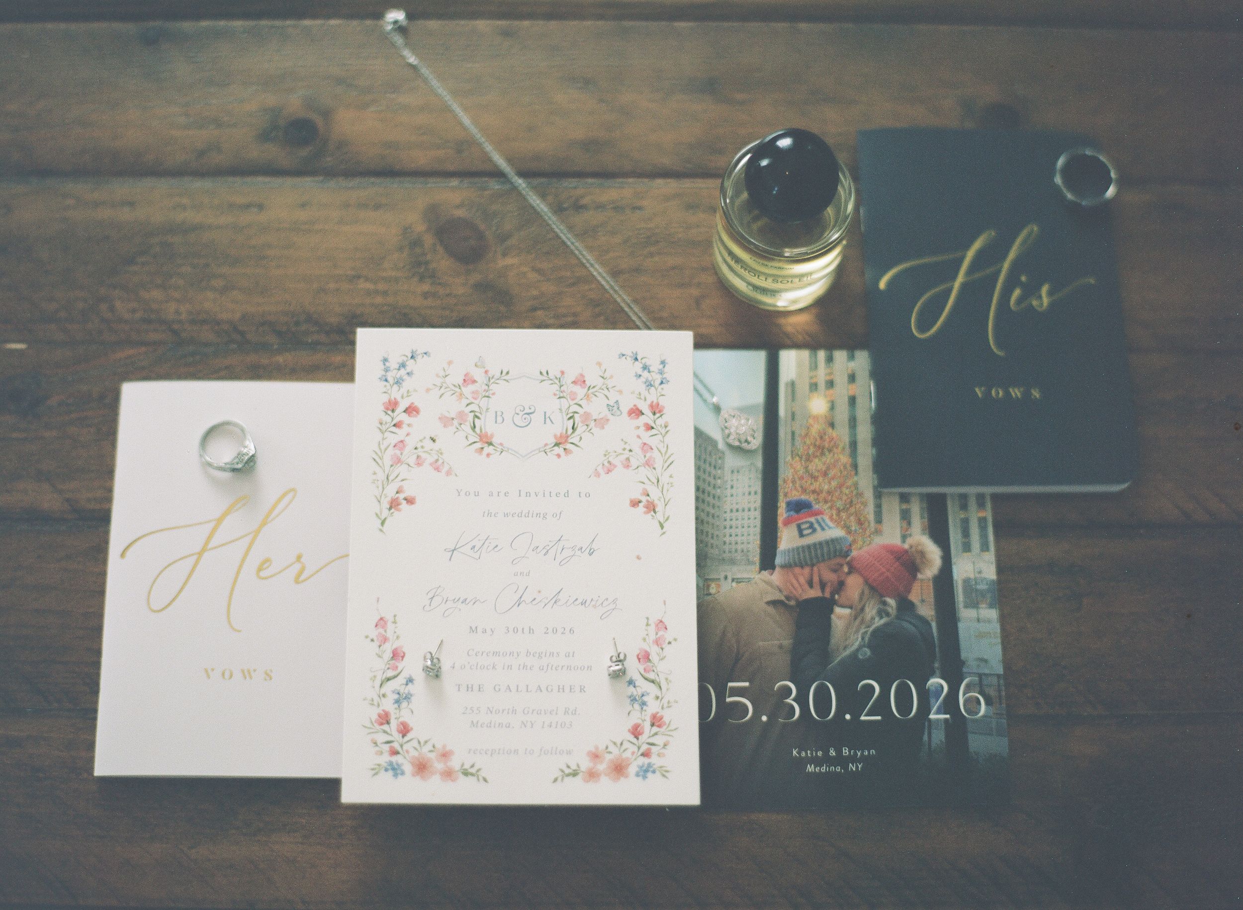 Wedding stationery flatlay with floral invitations, vow books, and rings on wood table.