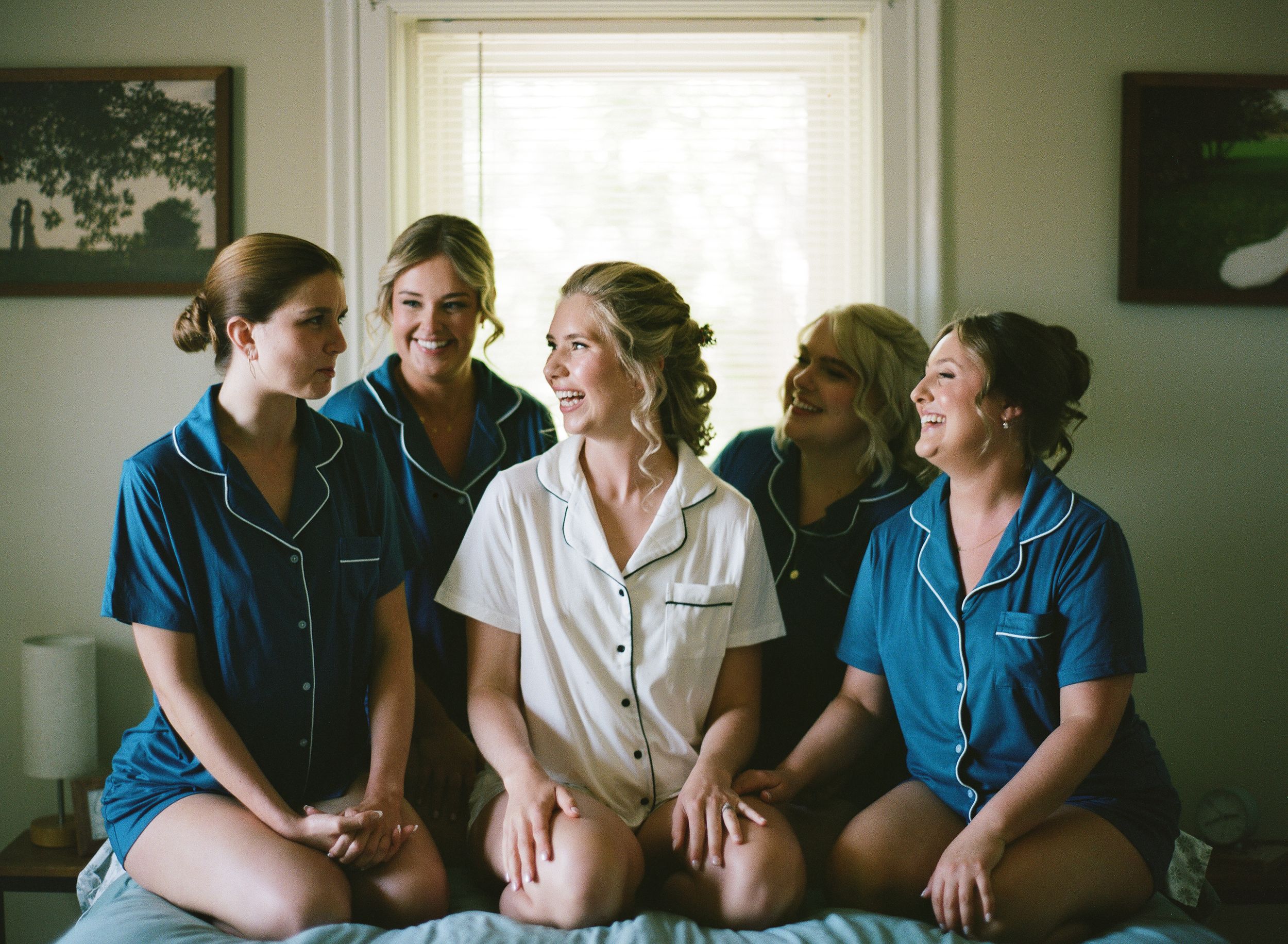 Bride and bridesmaids in matching blue and white pajamas sitting together on a bed, smiling and laughing.