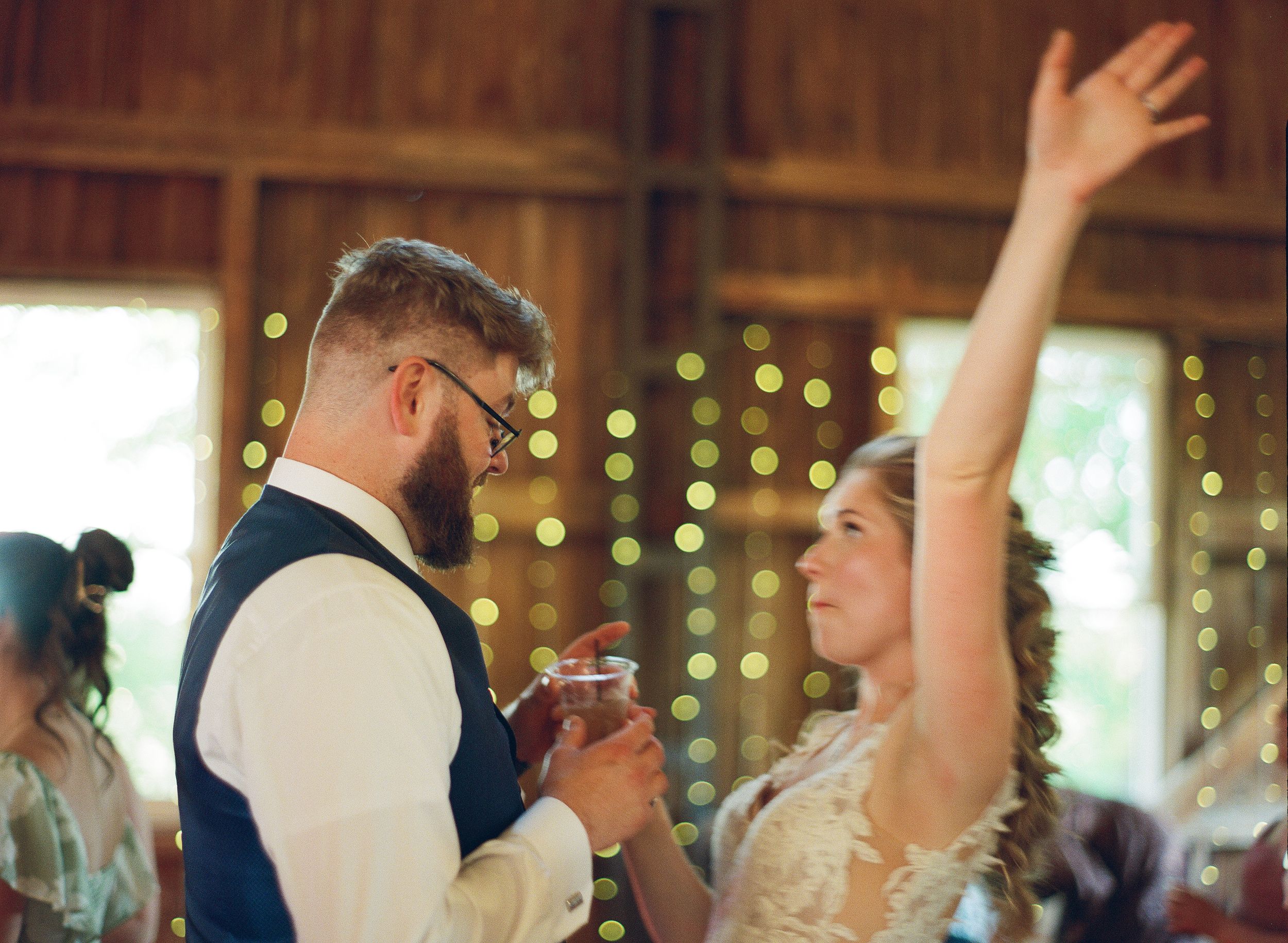 Bride raising her arm joyfully while dancing with groom in a rustic barn with string lights.