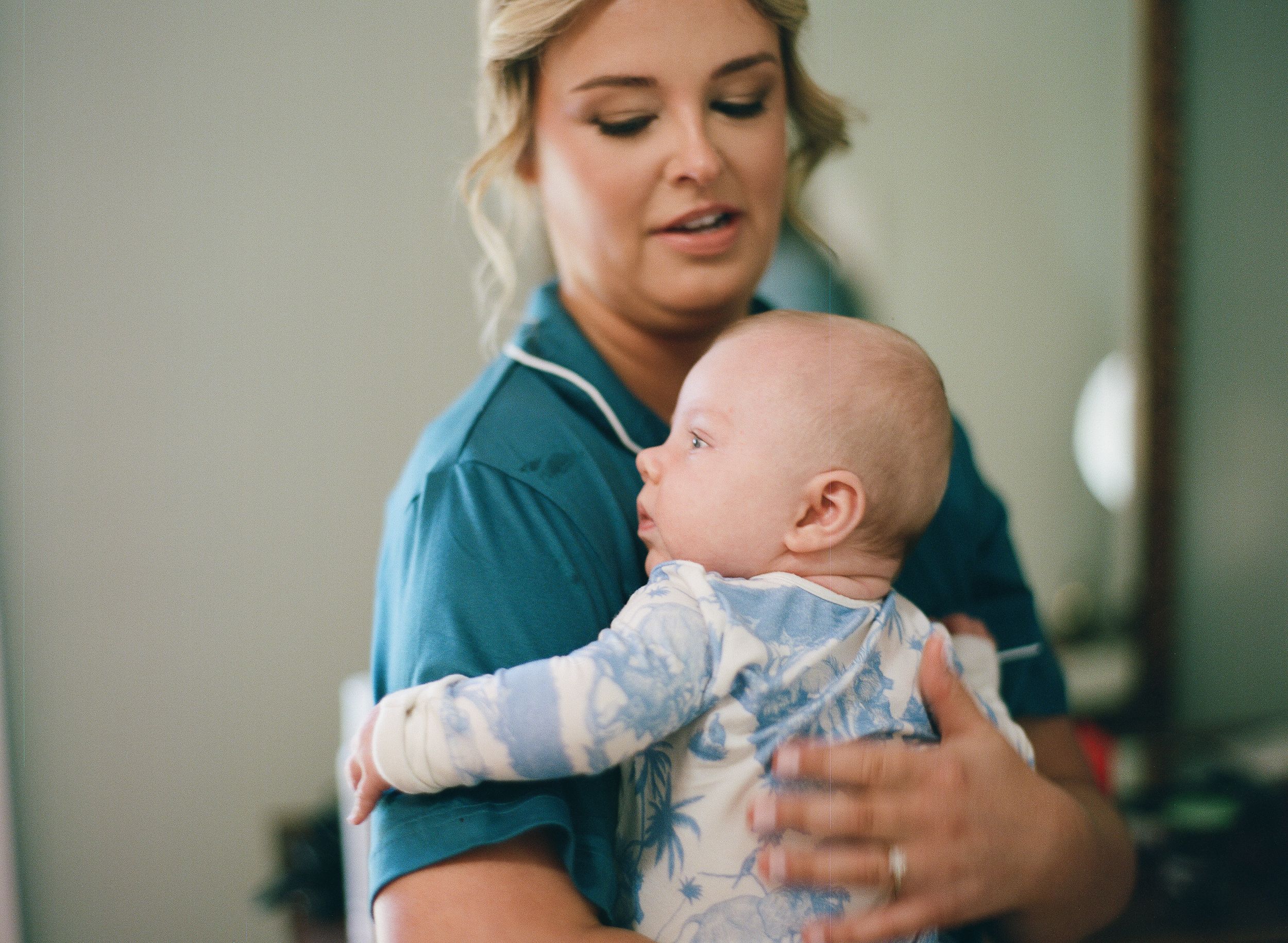 A nurse in teal scrubs gently holds a young baby in a light blue outfit in a clinical setting.