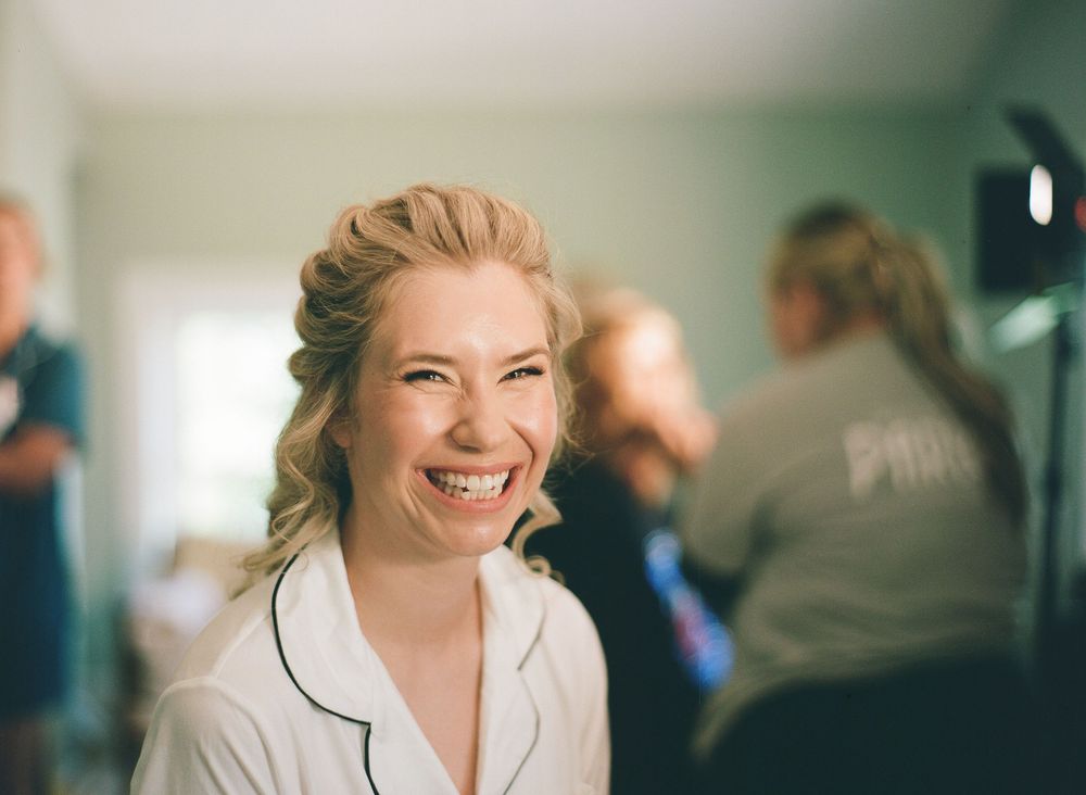 Smiling blonde bride in white robe laughing joyfully during wedding preparations with bridesmaids in background.