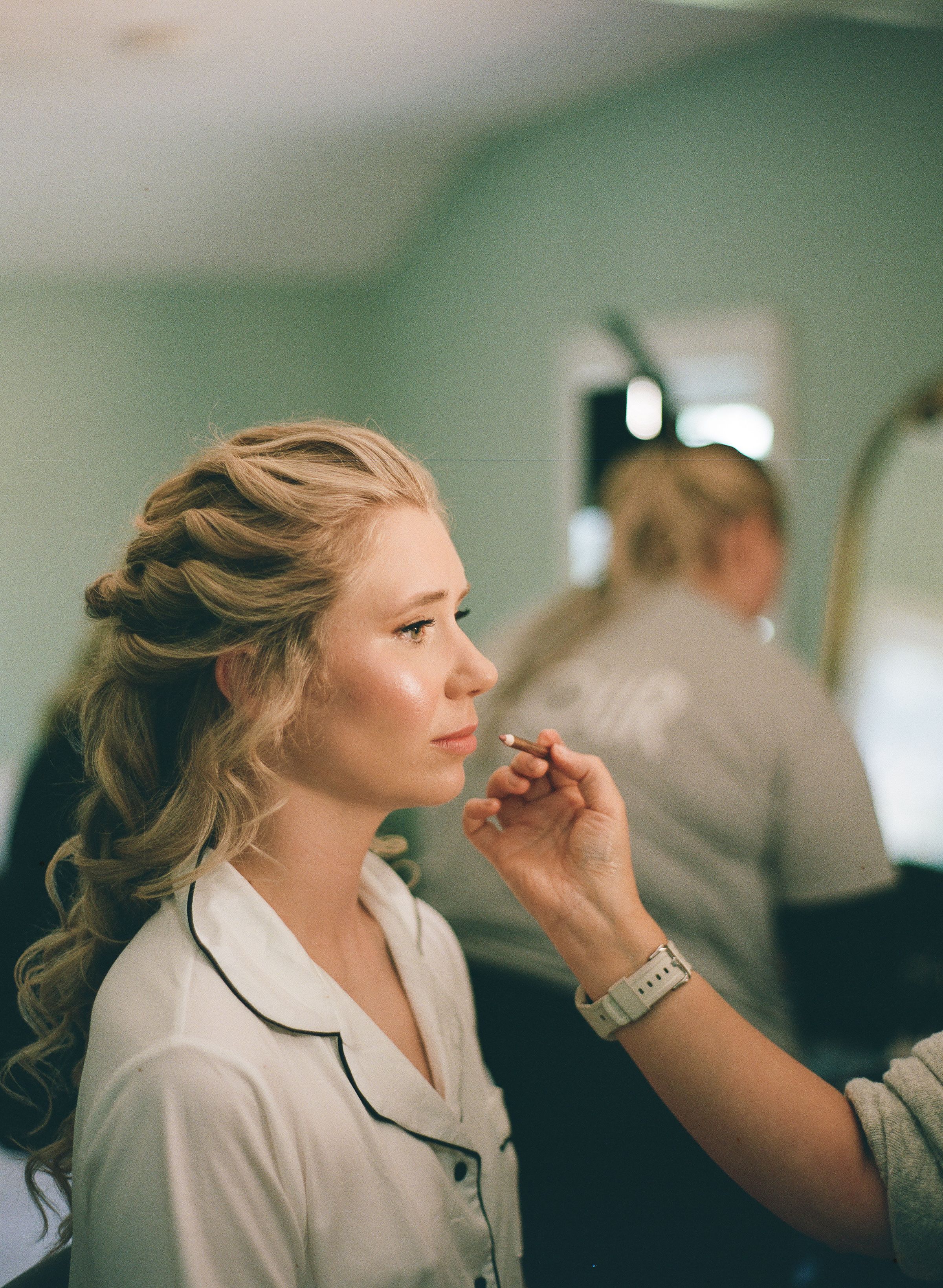 Bride with elegant updo gets makeup applied by artist in bright dressing room.