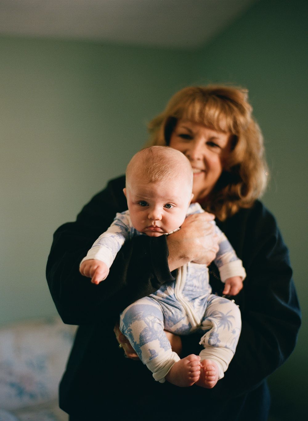 A smiling woman with blonde hair holds a baby in a blue floral onesie in a cozy indoor setting.